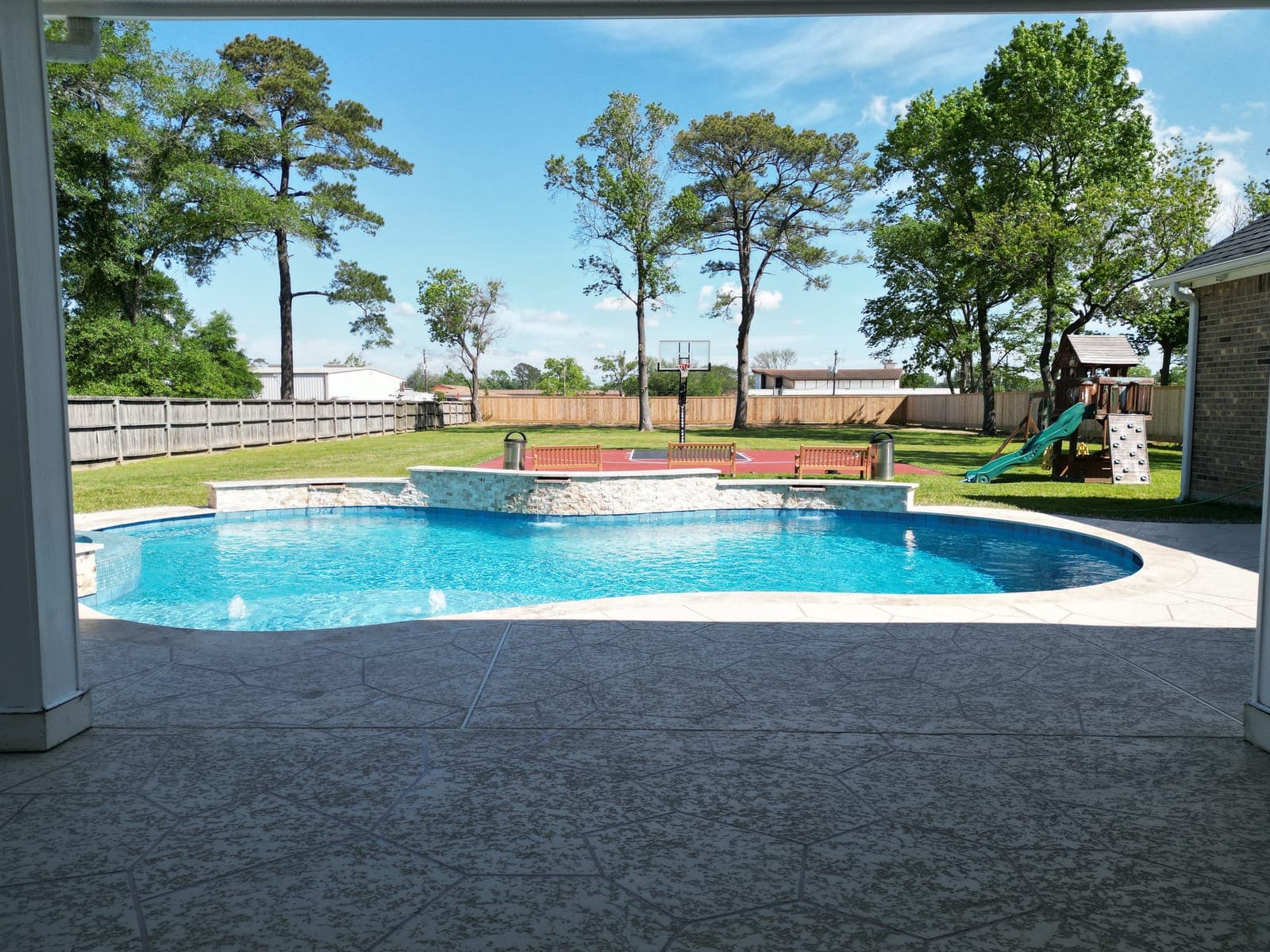 Custom pool in Highlands, TX. Framed from the covered patio: a freeform pool below a stacked-stone waterfall wall, with a sport court and a playset on the lawn behind it.