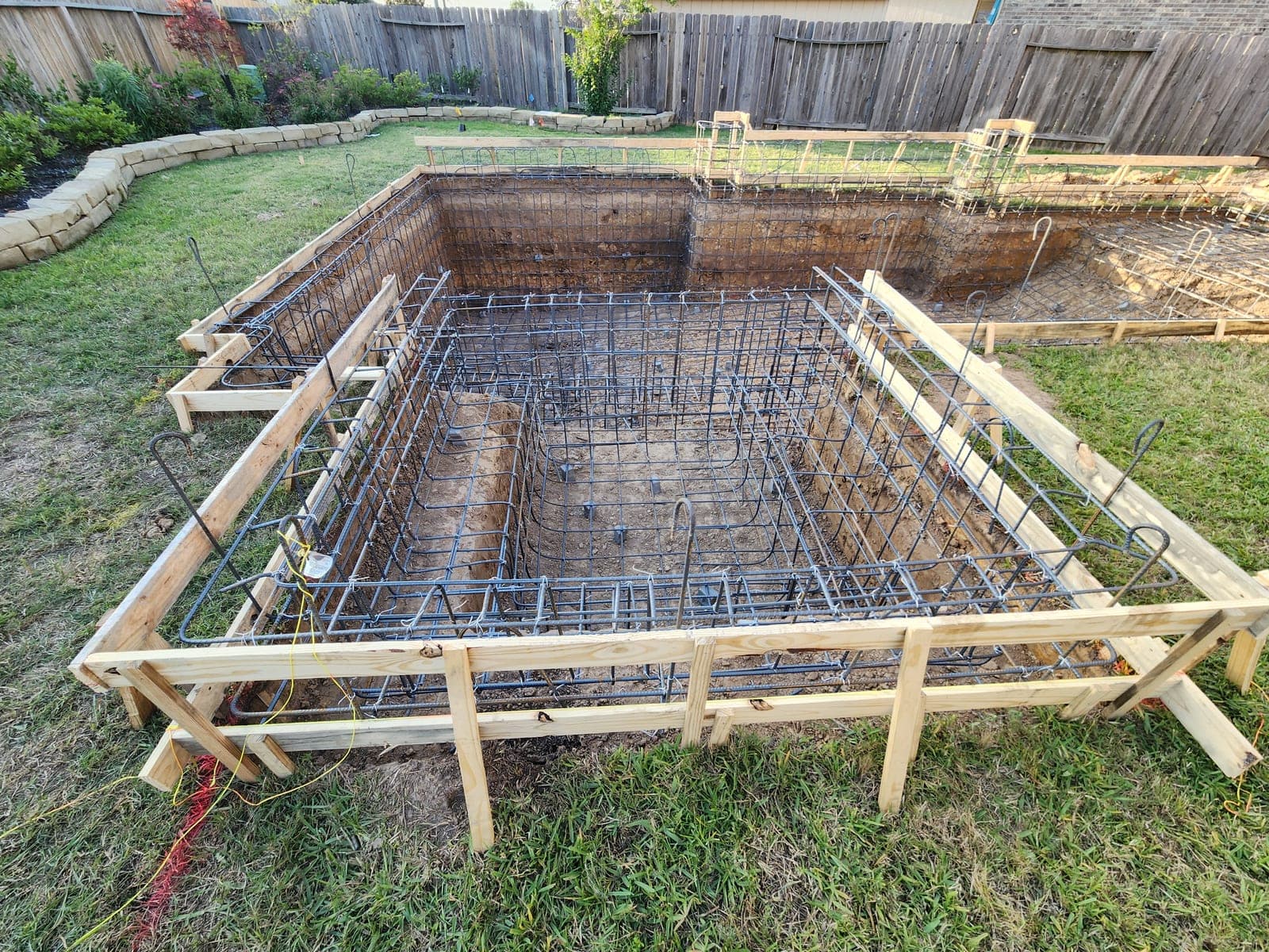Piscina custom en Spring, TX. Una excavación de piscina de forma libre, amarrada con una jaula completa de acero, tablas de formaleta en el borde, un macizo de piedra apilada y una cerca privada detrás.