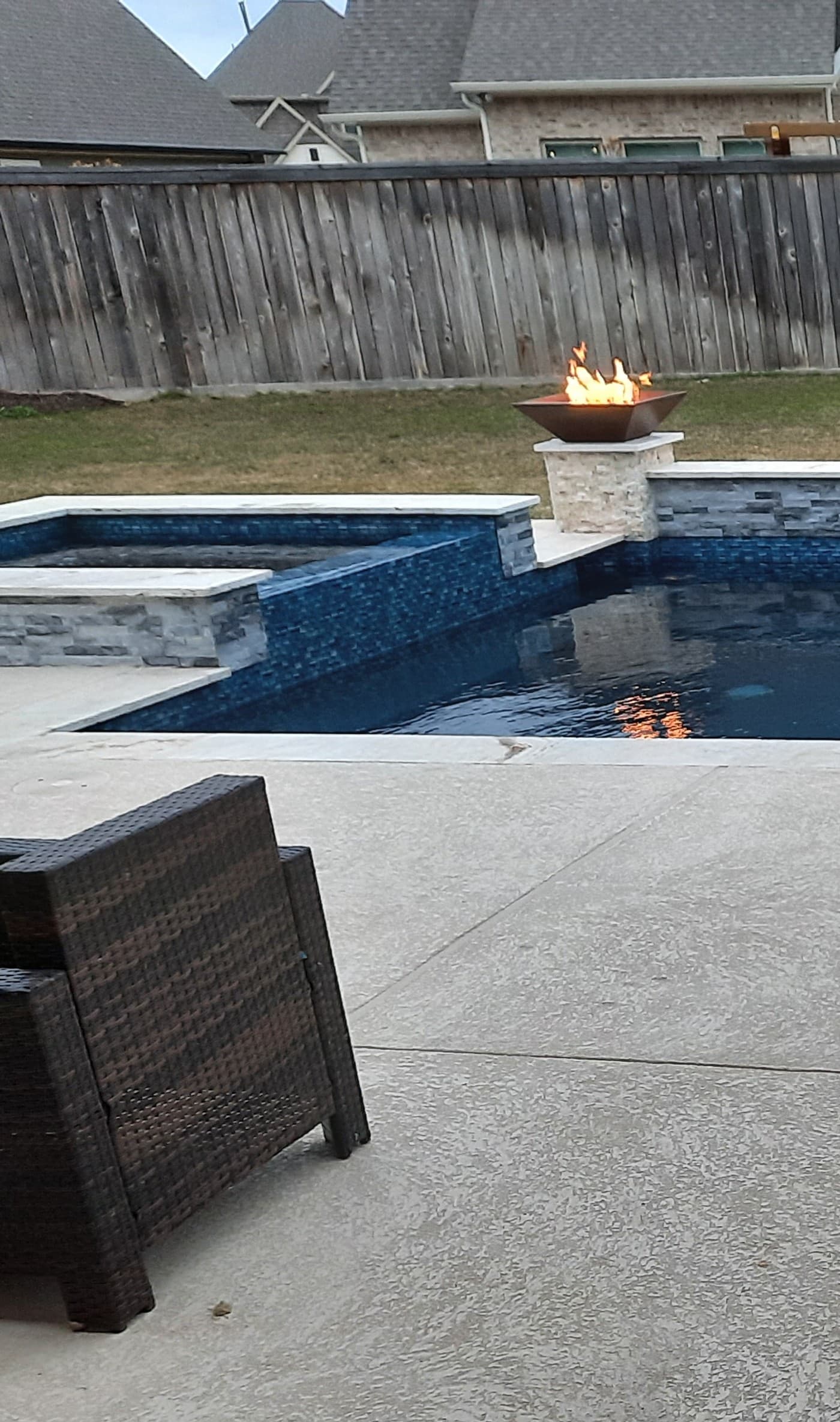 Piscina custom en Bridgeland, TX. Un spa elevado revestido en mosaico azul, un fogonero encendido sobre un pedestal de piedra justo detrás del muro, y una silla de mimbre oscura esperando en el patio en primer plano.