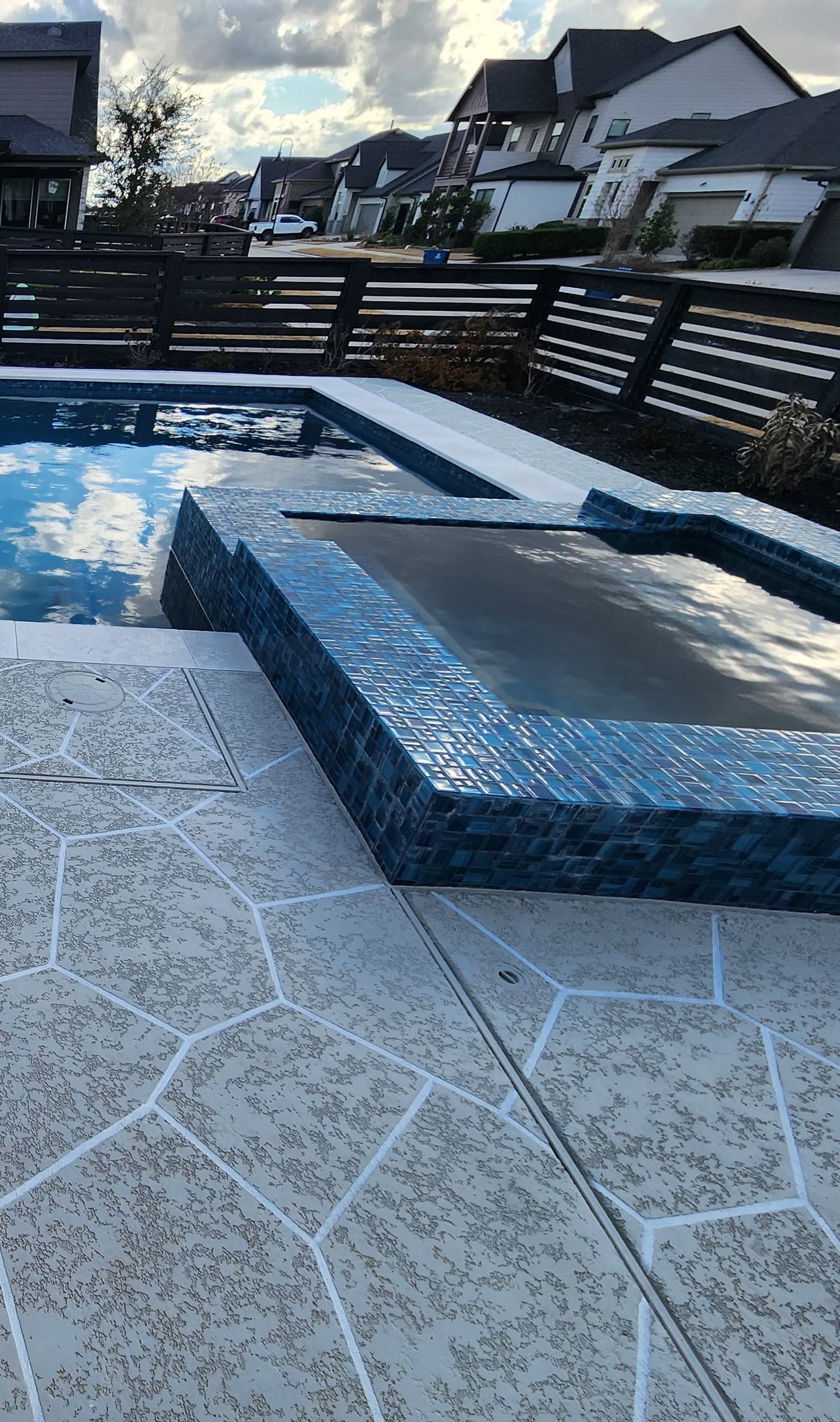 Piscina custom en Bridgeland, TX. Un muro de spa en mosaico de vidrio azul se une a una esquina de deck con patrón hexagonal, el agua oscura de la piscina reflejando las nubes y los techos vecinos detrás de la cerca privada.
