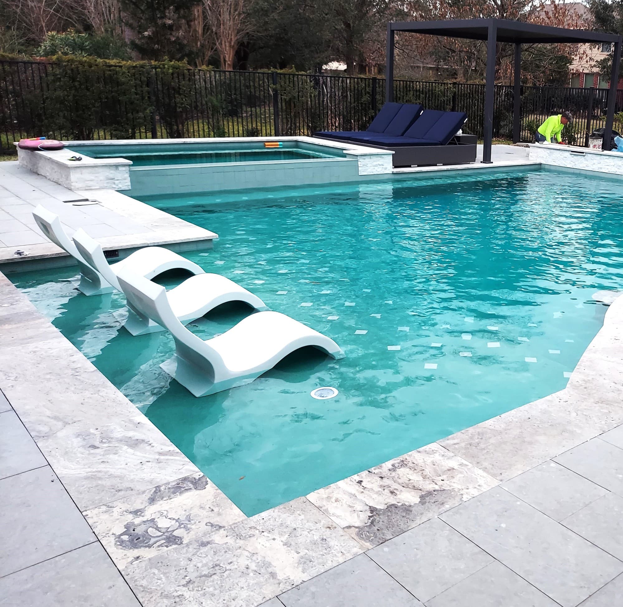 Piscina custom en Bridgeland, TX. Una vista elevada de la misma piscina: tres camastros blancos ondulados flotan en la zona baja, un spa rectangular elevado en travertino se asienta en la esquina lejana, y una pérgola negra cubre camastros con cojines azul marino junto a una verja de hierro forjado y árboles maduros.