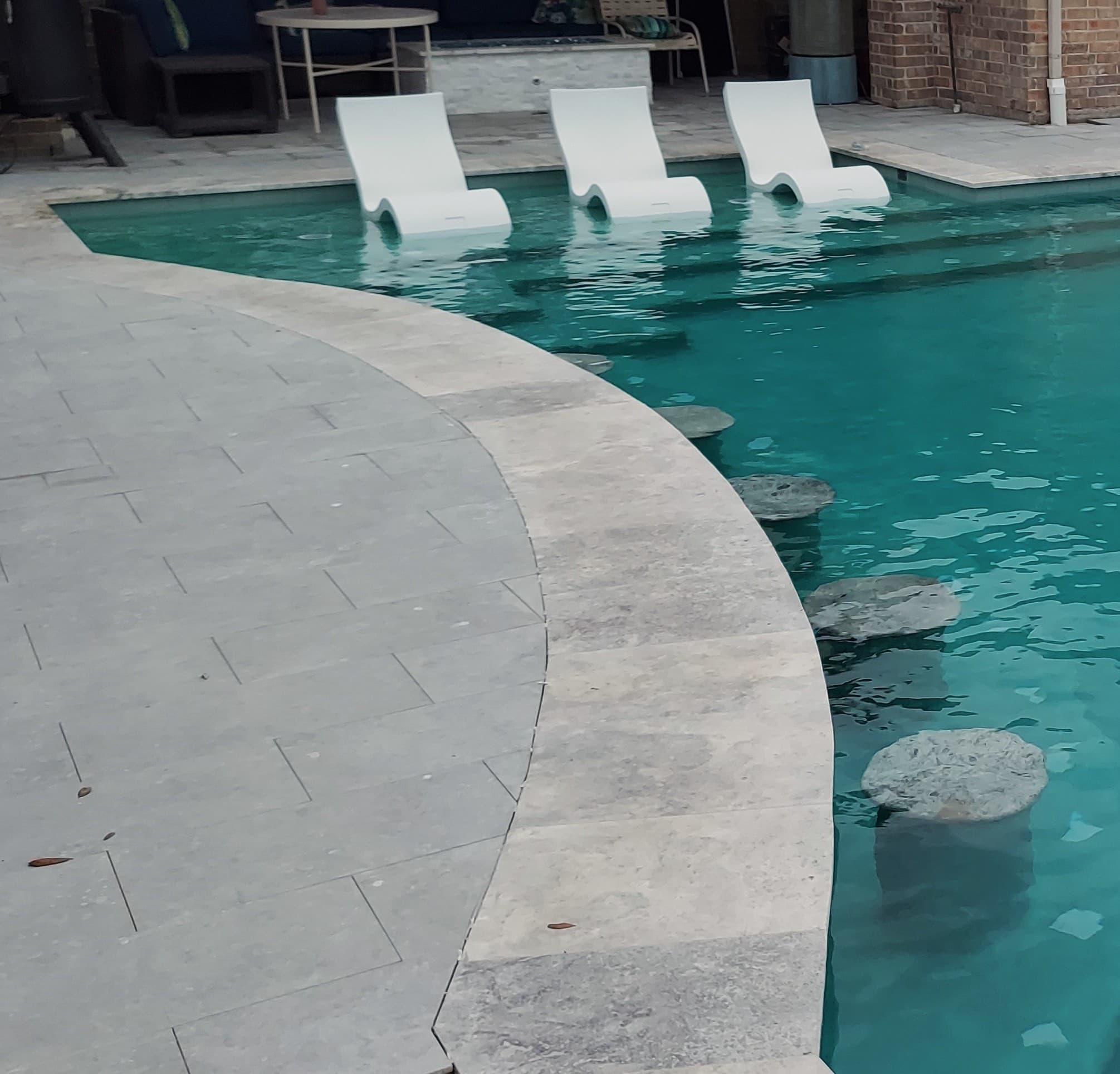 Piscina custom en Bridgeland, TX. Un borde de piscina curvo en travertino con tres camastros blancos flotando en la zona baja, amplias piedras pisables dentro del agua turquesa que llevan hacia un patio cubierto con una mesa de fuego en piedra y un muro de ladrillo detrás.