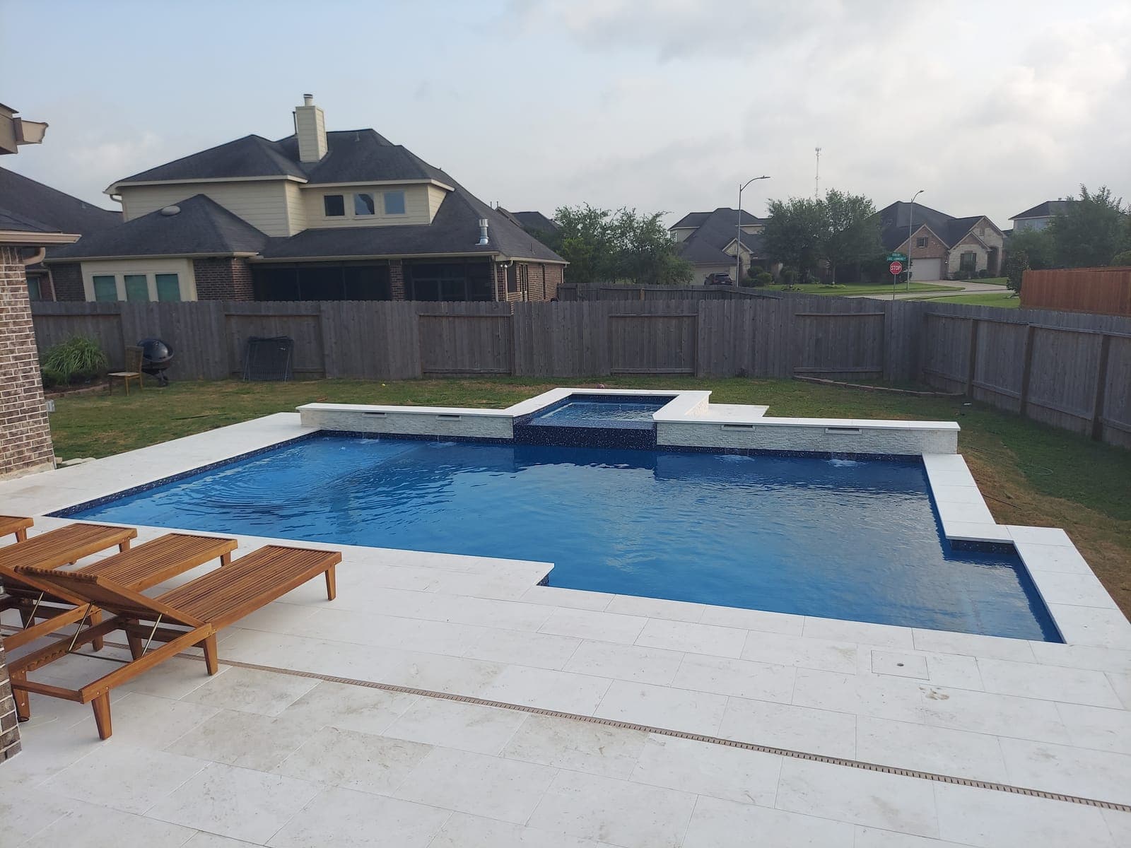 Piscina custom en Rosenberg, TX. Bajo un cielo gris parejo el acabado oscuro se lee azul profundo: dos muros elevados vierten agua por sus vertedores, el spa queda entre ellos y una franja de azulejo cruza el deck de travertino claro.