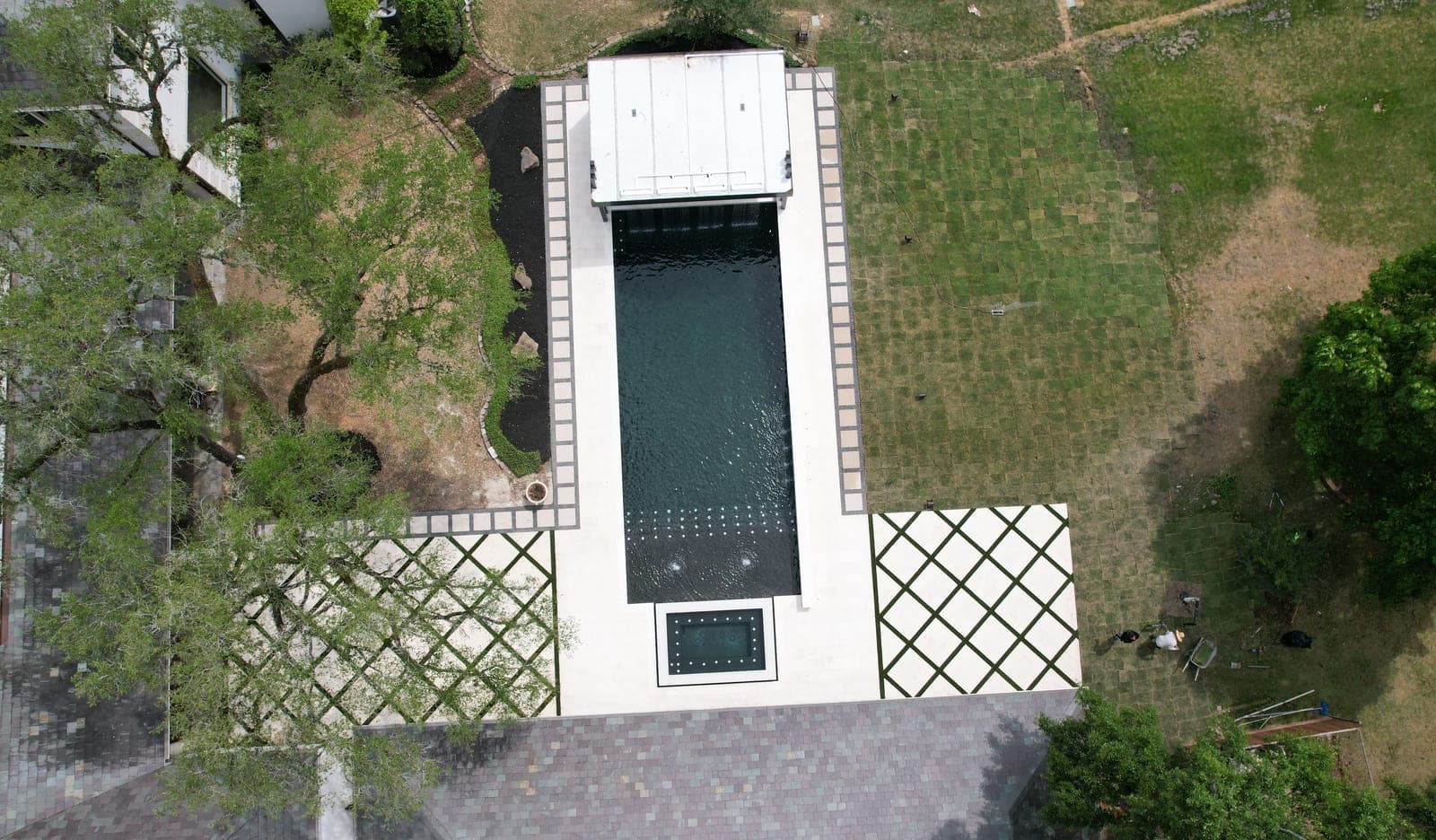 Piscina de concreto en Houston, TX. A plomo sobre agua oscura: una lámina cae desde una cabaña revestida en piedra en un extremo, un deck de patrón diamante bordeado de césped corre al lado, y un spa elevado con chorros se asienta justo pasado el borde lejano.
