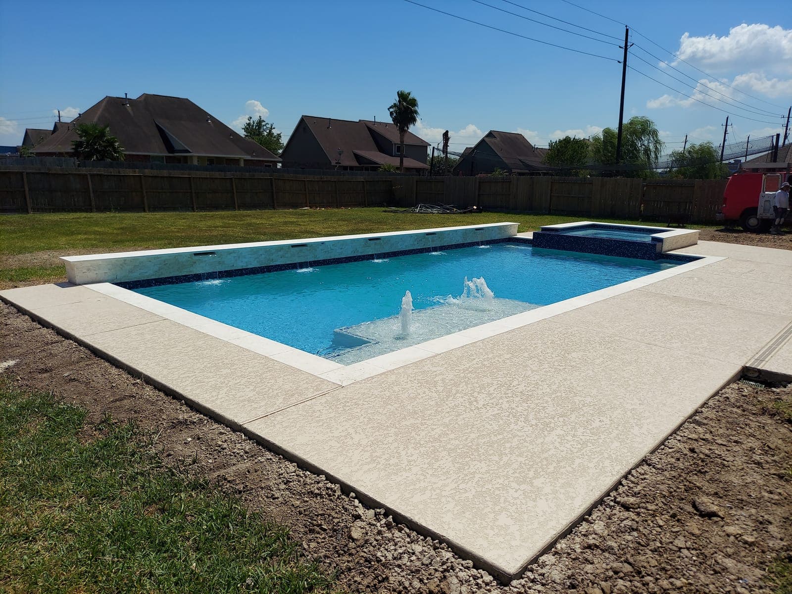 Piscina de concreto en Mont Belvieu, TX. A días de terminarse, con la tierra aún desnuda al borde del deck: el agua cae en lámina desde un muro de azulejo azul marino, los burbujeadores se levantan del banco solar y el spa elevado viste mosaico oscuro.