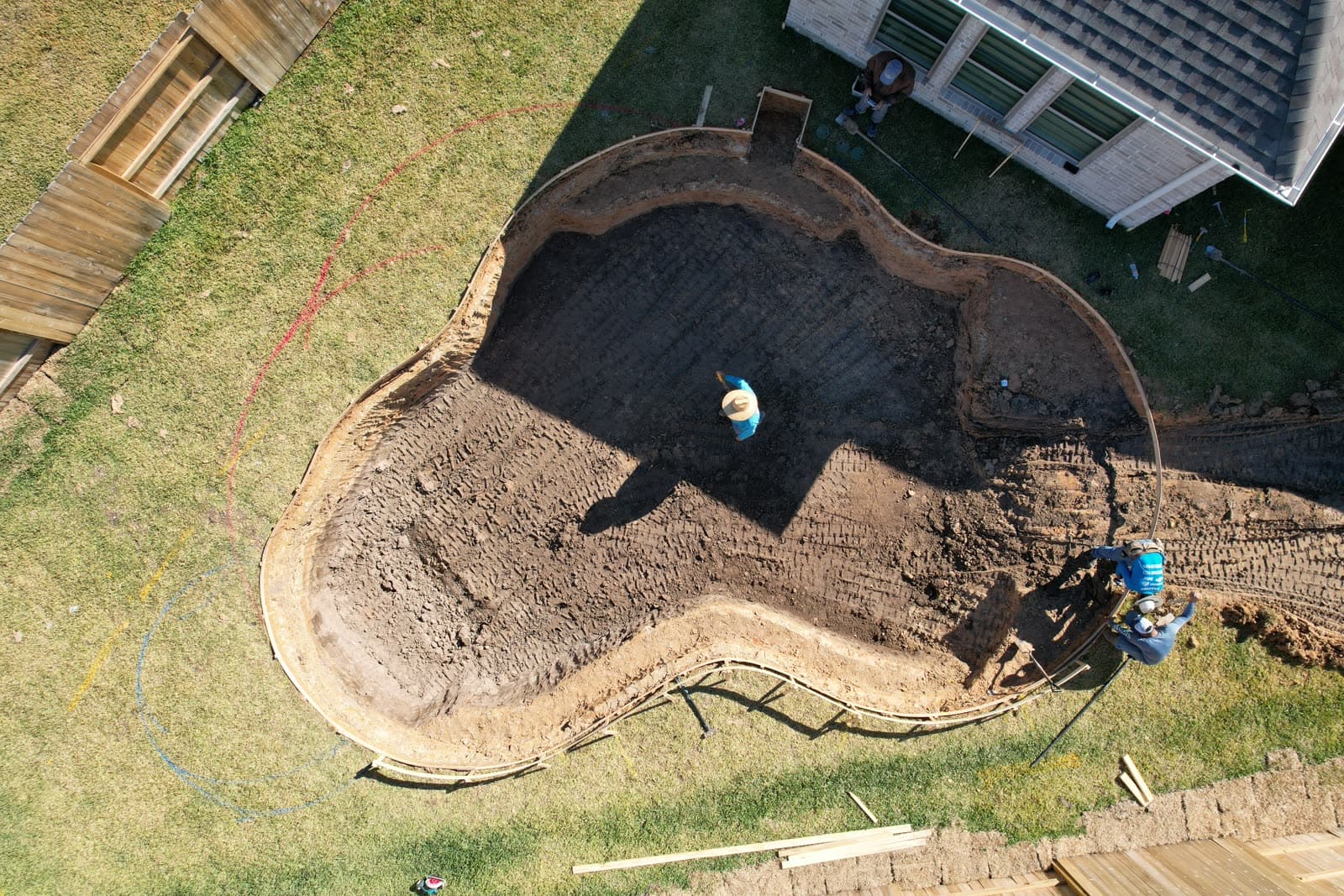 Piscina custom en Cypress, TX. A plomo sobre una excavación recién hecha: la forma libre cortada en la tierra cruda de Houston, las líneas de replanteo aún pintadas sobre el césped y un obrero de pie dentro.