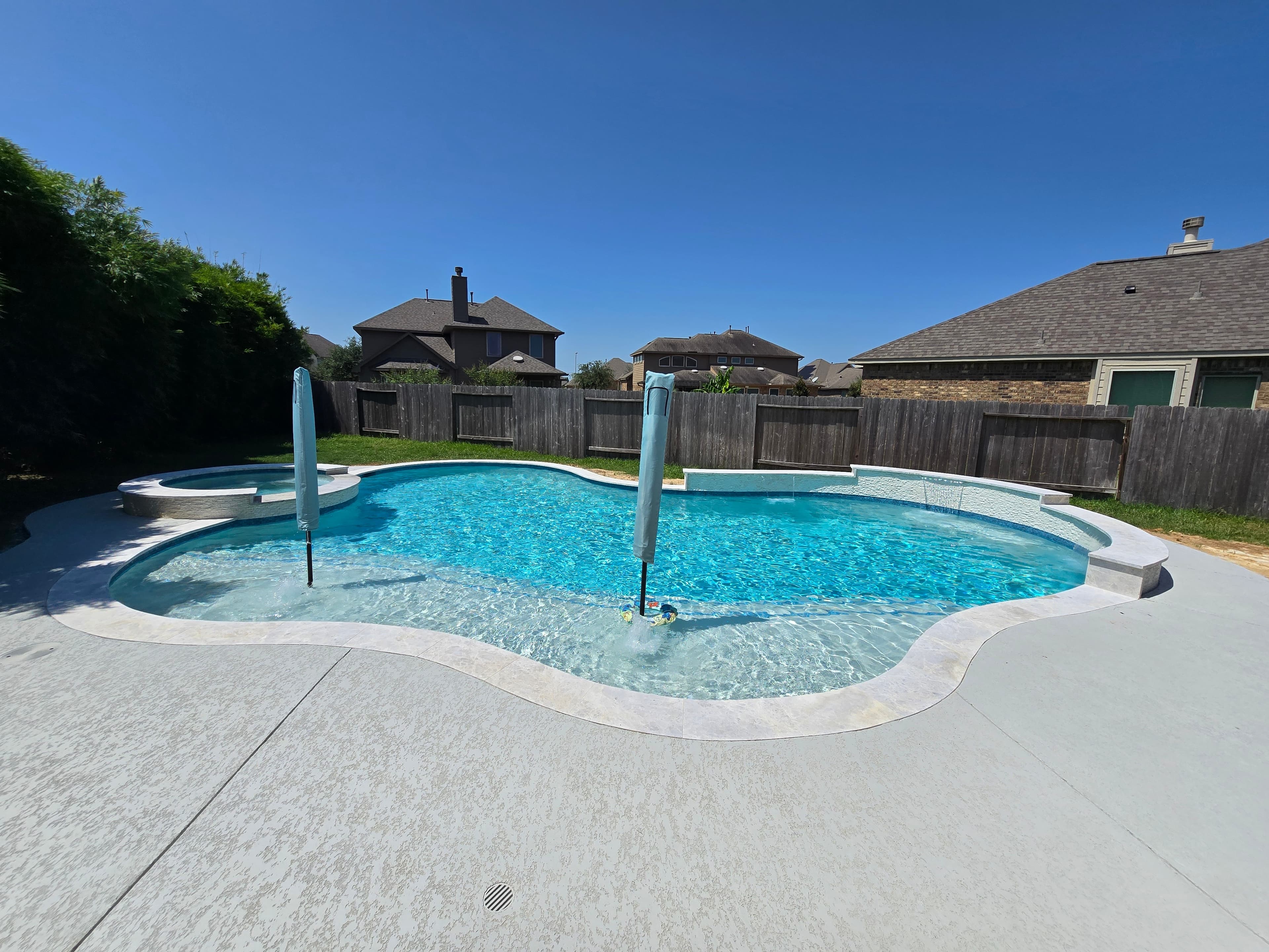 Piscina custom en Hockley, TX. Una piscina de forma libre con spa redondo elevado, una cascada de lámina que vierte hacia ella, y fundas de sombrilla azules en el deck.