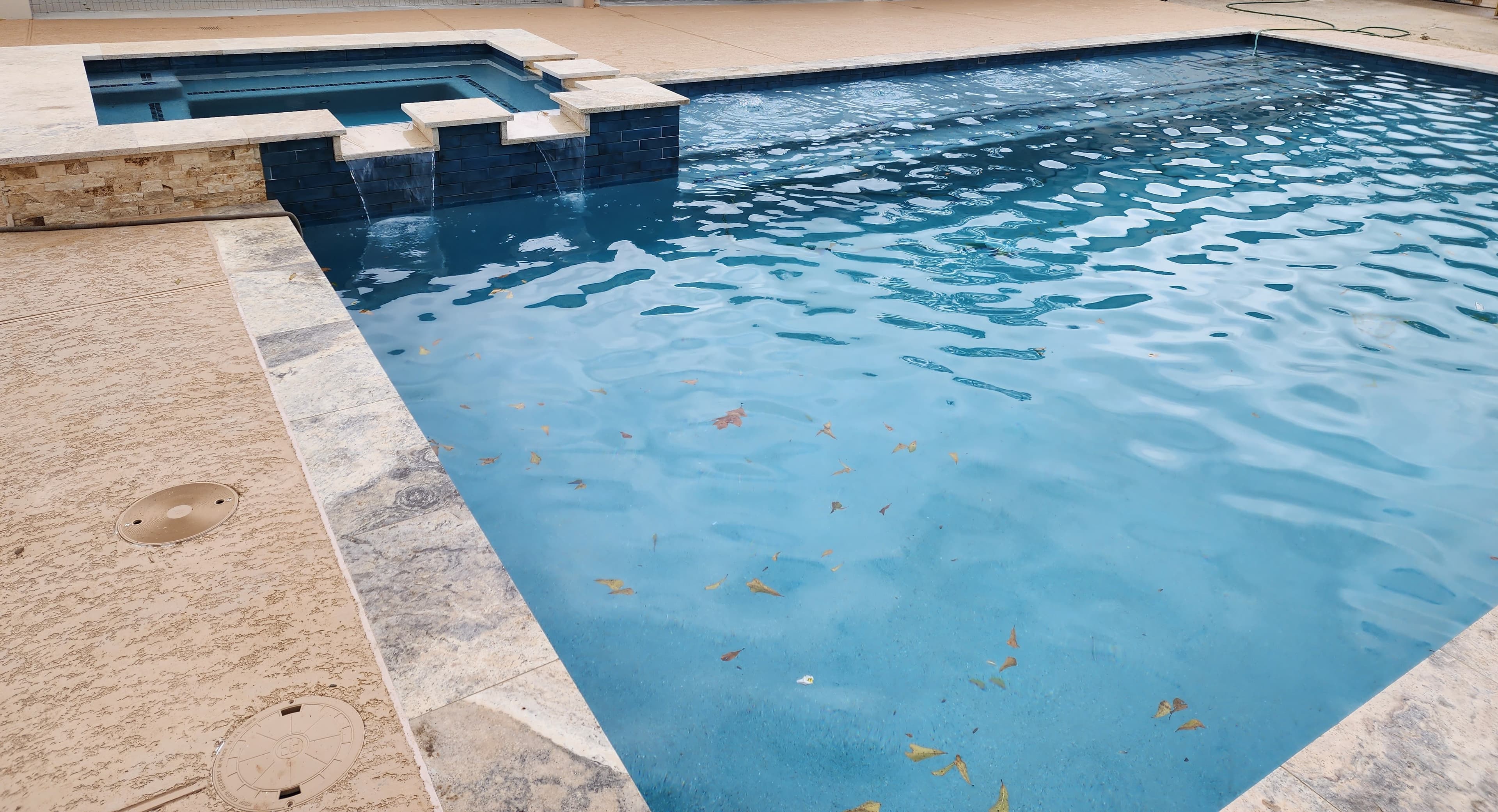 Piscina custom en Huffman, TX. Una piscina rectangular se une a un spa elevado revestido en mosaico de vidrio iridiscente, con el agua cayendo en lámina de cascada desde su muro de azulejo oscuro, y el coping de travertino recorriendo el borde a pleno sol.