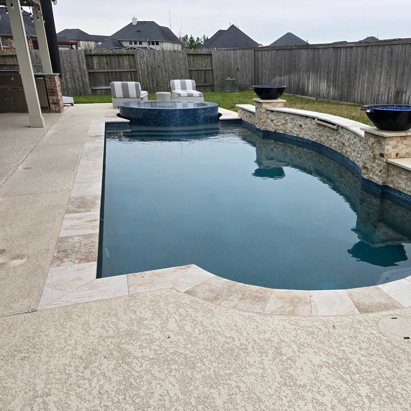 Custom pool in Katy, TX. Two black fire bowls sit on stone pedestals above a curved stacked-stone wall, with a round mosaic spa raised at the far end.