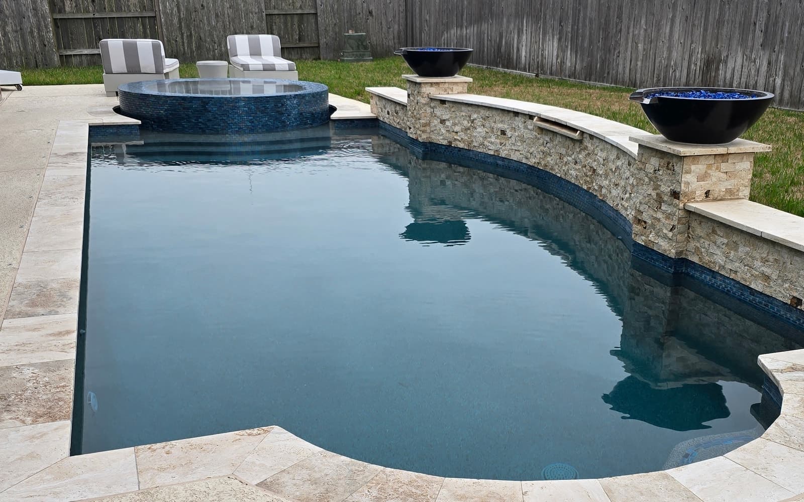 Piscina custom en Katy, TX. Un spa redondo elevado en mosaico de vidrio azul junto a la piscina, con dos fogoneros negros ardiendo sobre un muro curvo de piedra apilada que recorre el patio.