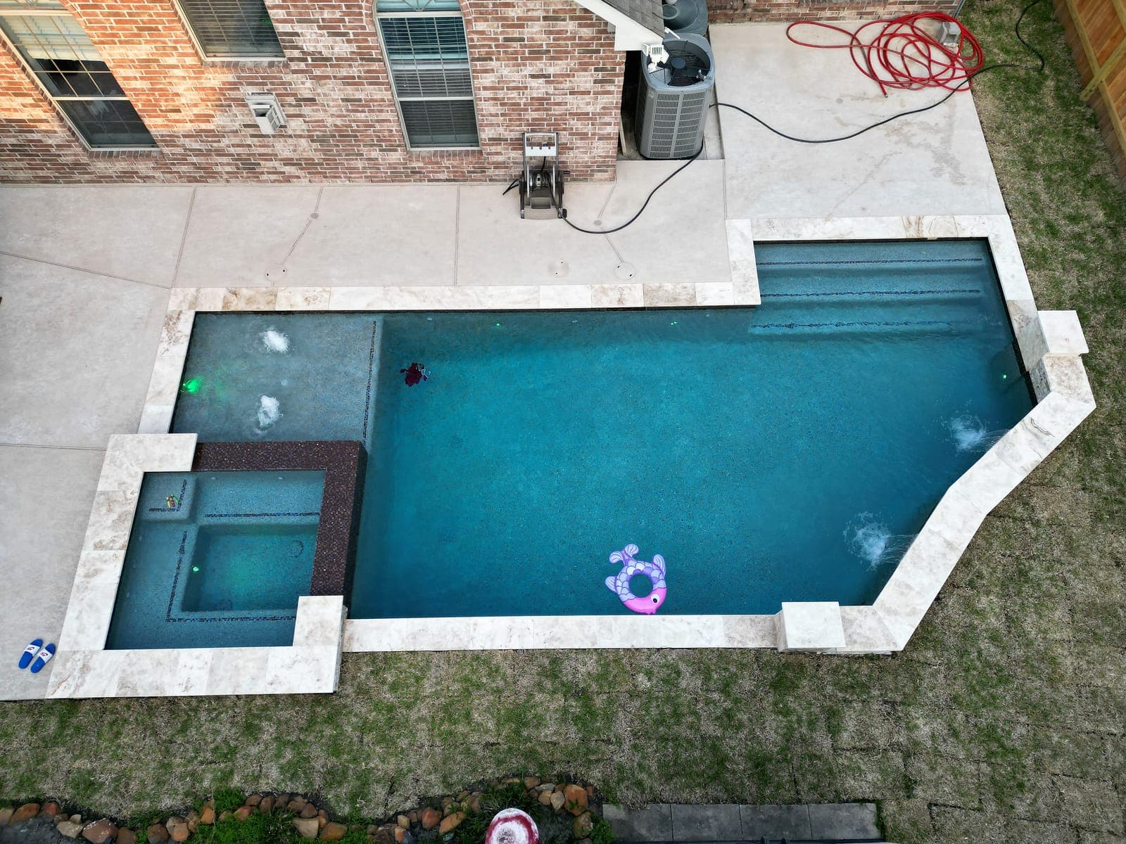 Custom pool in League City, TX. Straight down at dusk: the underwater lights just on, air jets churning white at the corners, and the raised spa glowing green inside its dark mosaic band.