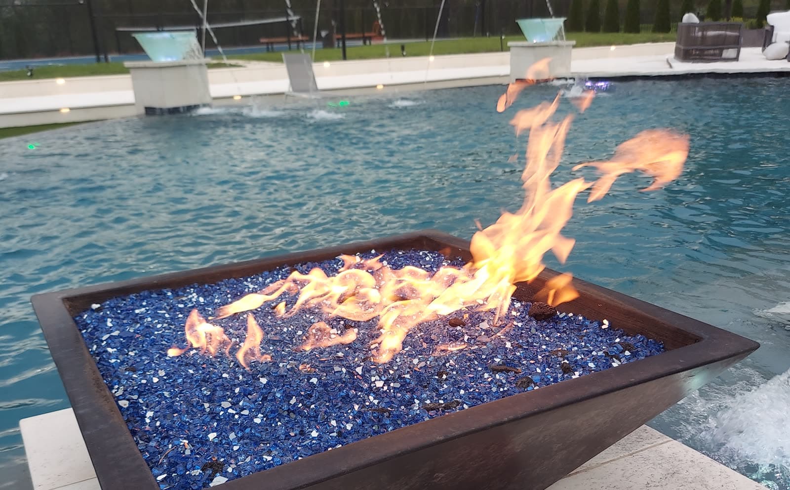 Custom pool in Richmond, TX. A copper fire bowl burning over blue fire glass, set in the water at dusk with fountain jets behind it.