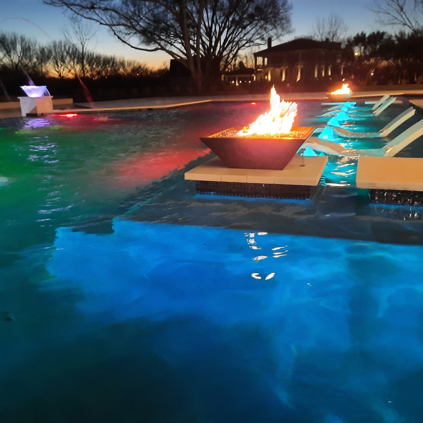 Custom pool in Richmond, TX. Blue hour, everything on: a fire bowl burning in the middle of the water, lit jets arcing over the surface, the loungers glowing from below and the pool floor turned electric blue.