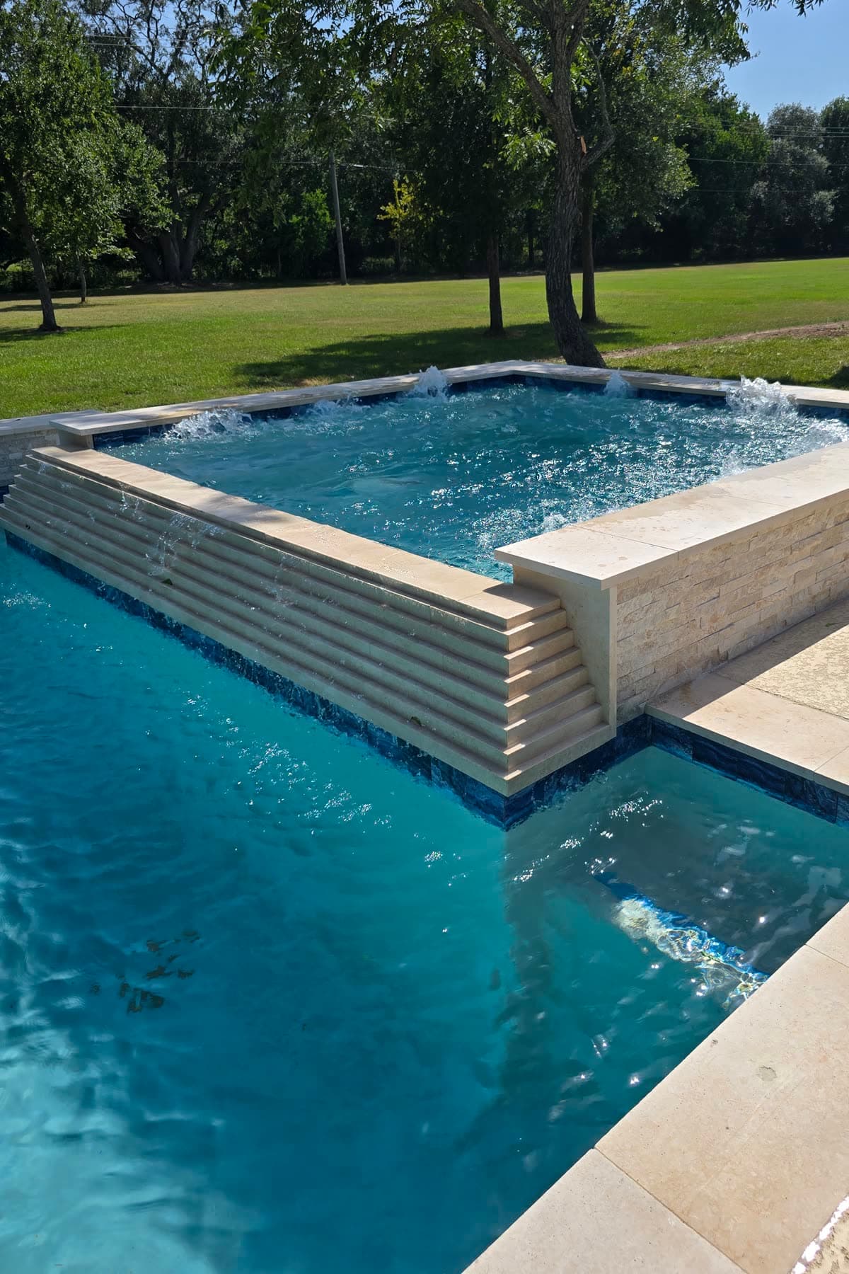Custom pool in Richmond, TX. A raised spa faced in stacked stone, water sheeting off its wall into the pool below, jets running.
