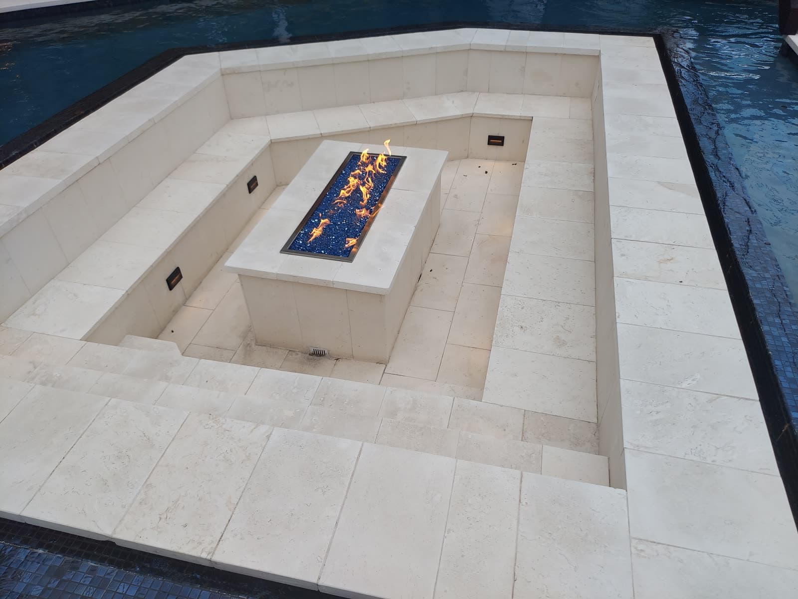 Piscina custom en Richmond, TX. Una sala hundida dentro de la piscina, cerrada en travertino, con una línea de fuego encendida sobre vidrio azul en el centro.