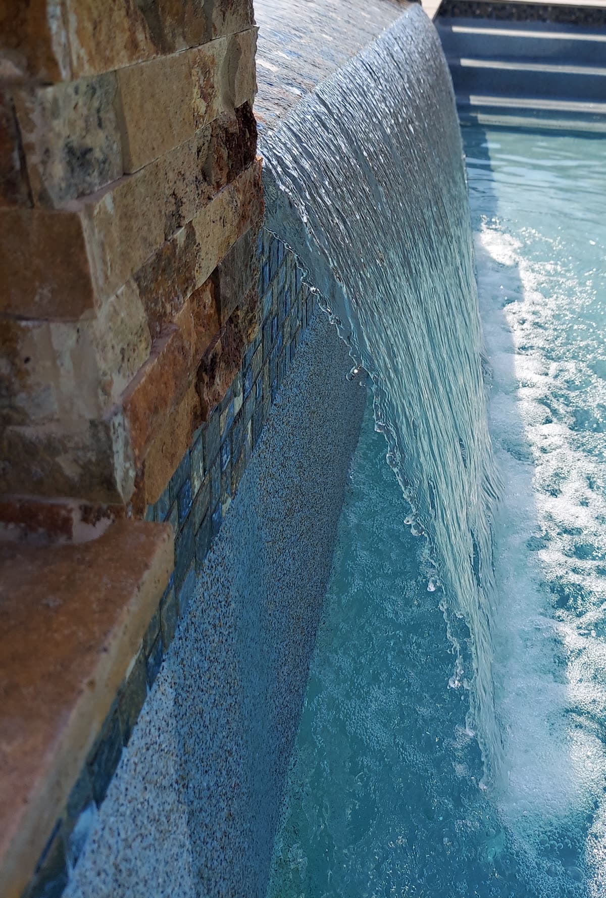Custom pool in Tomball, TX. Close on a sheer descent: water curves over stacked stone and glass tile and lands in a line of bubbles.