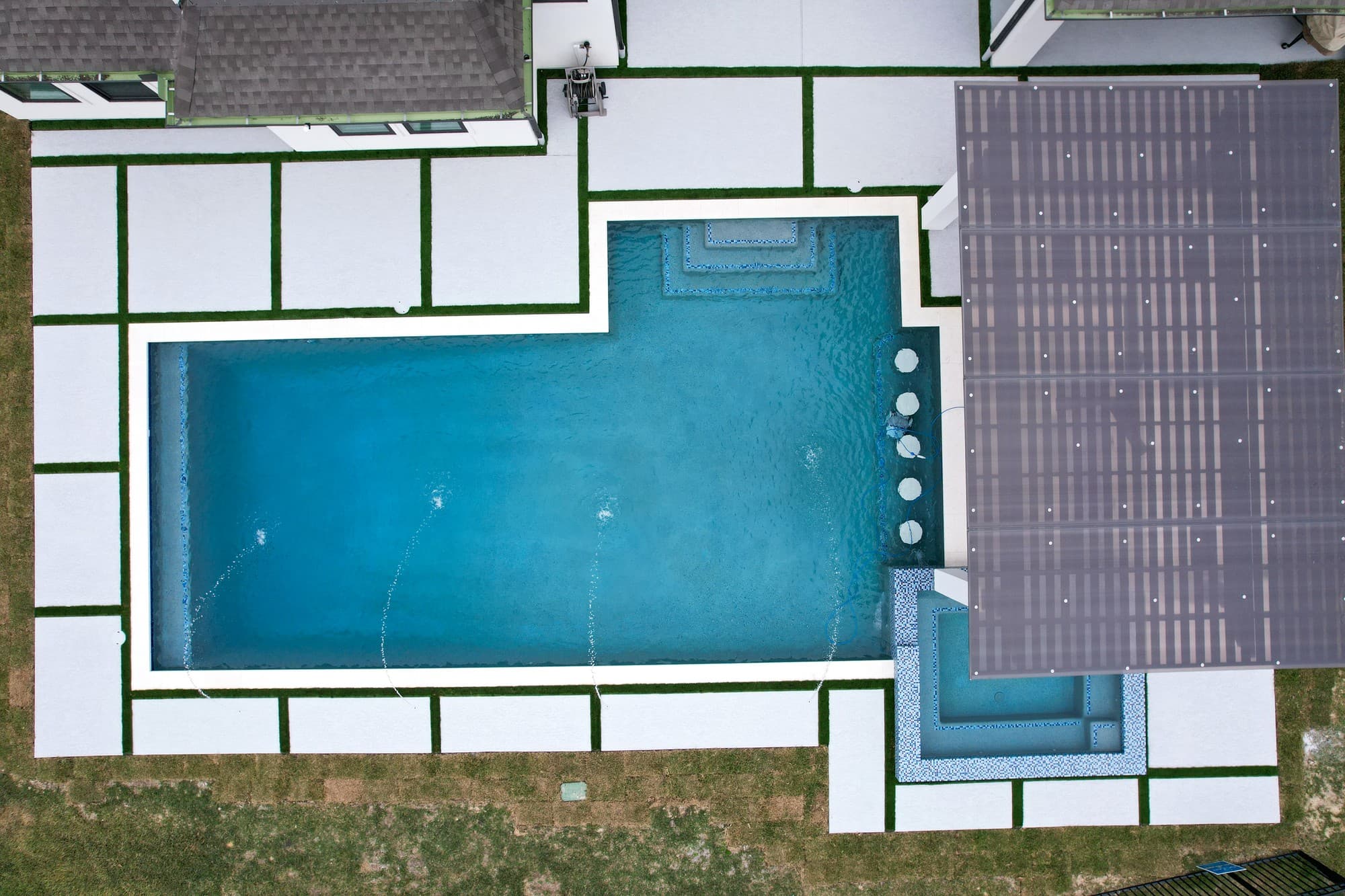 Custom pool in Cypress, TX. Seen straight down: a geometric pool with two square spas in blue mosaic at opposite corners, three deck jets arcing in, a row of in-water stools, a large pergola roof and a pale deck with turf strips between the pavers.