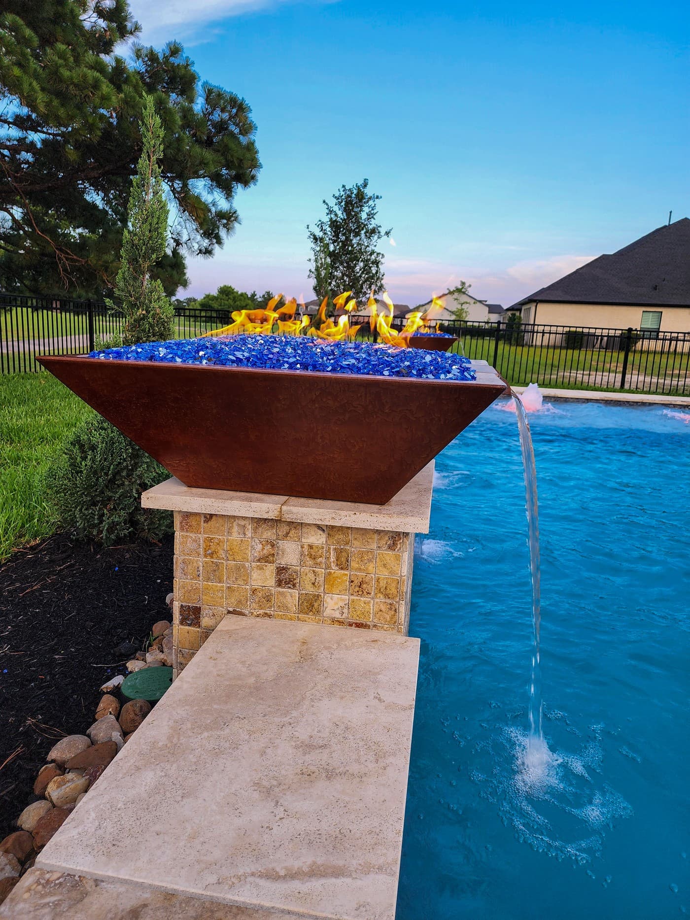 Custom pool in Cypress, TX. A copper fire bowl burning over blue fire glass, water pouring from its spout into the pool below, its pedestal faced in gold-tan mosaic tile.