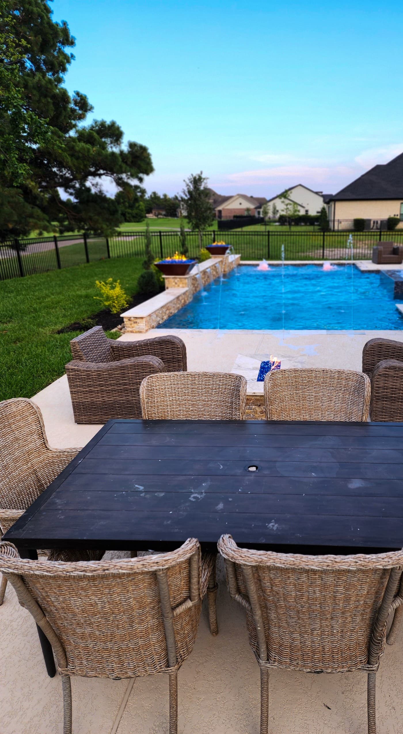 Custom pool in Cypress, TX. From the outdoor dining table, wicker chairs around it, toward the pool: deck jets in the air and two copper fire bowls burning on the raised wall at the far end.