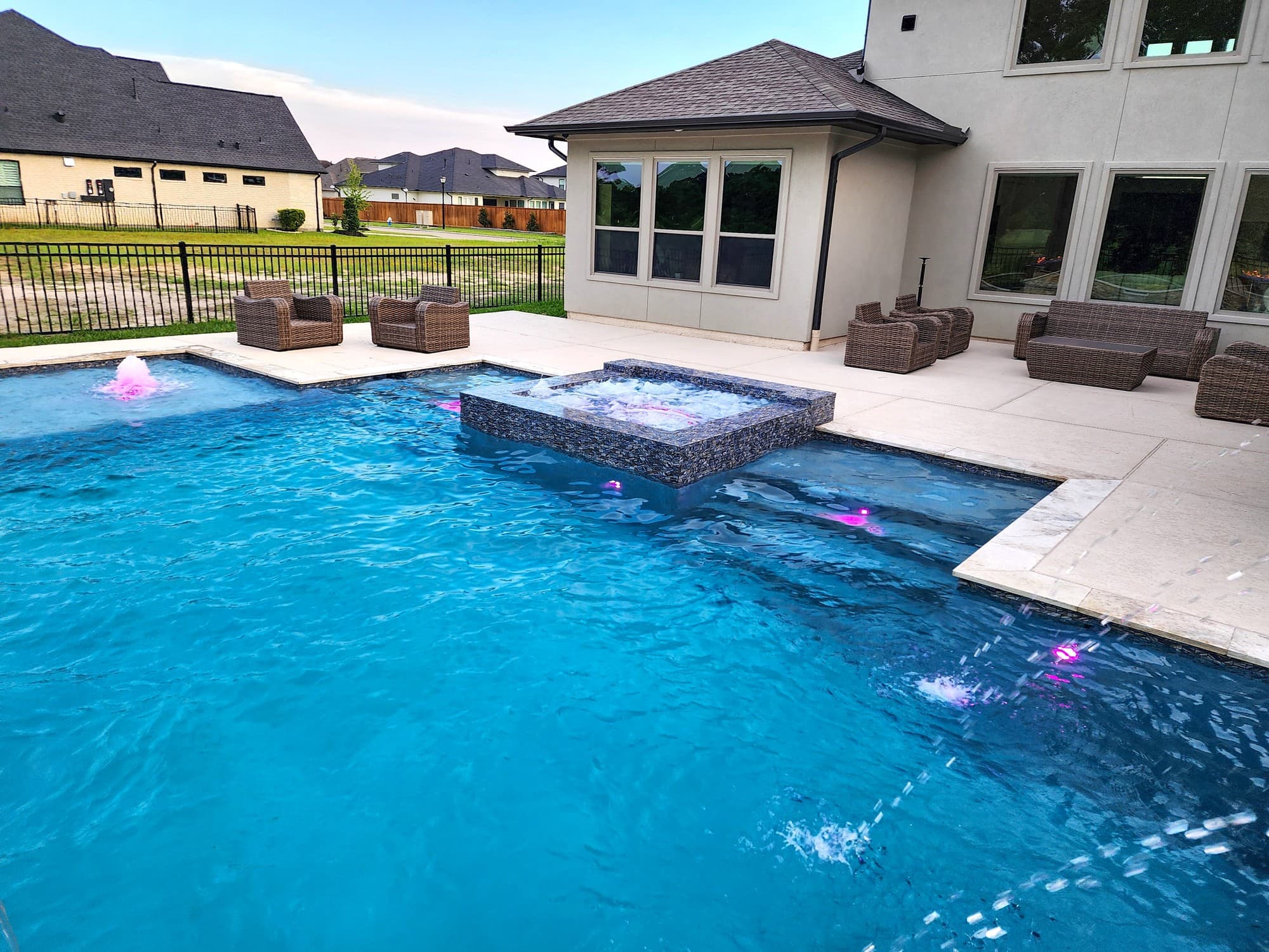Custom pool in Cypress, TX. At dusk, a geometric pool with a square raised spa clad in dark speckled stone spilling into it, deck jets arcing across the water, bubblers lit pink and wicker armchairs on the deck beside the house.