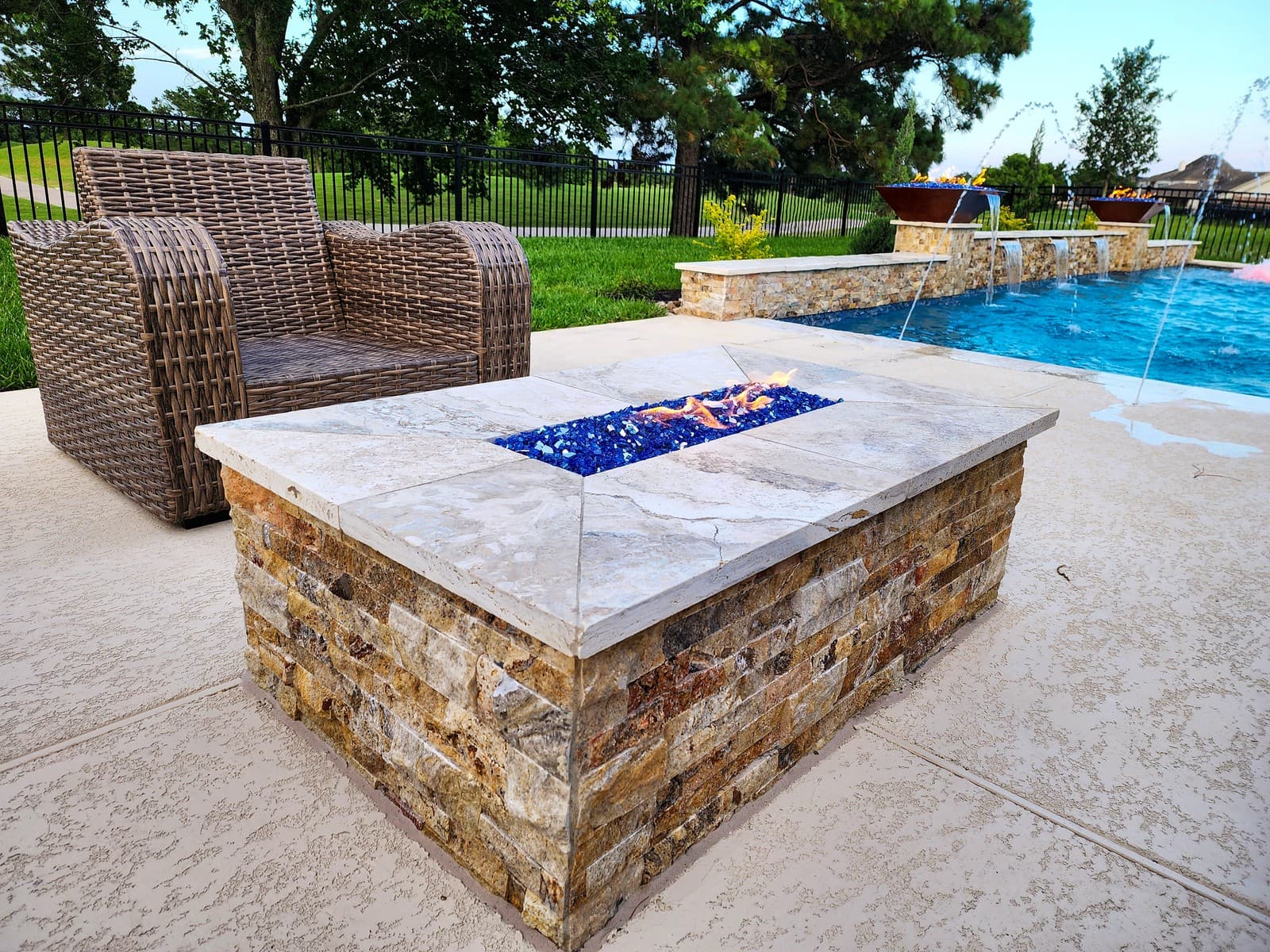 Custom pool in Cypress, TX. A rectangular stone fire pit on a stacked-stone base, blue fire glass burning under a stone top, a wicker armchair beside it and deck jets and fire bowls behind.