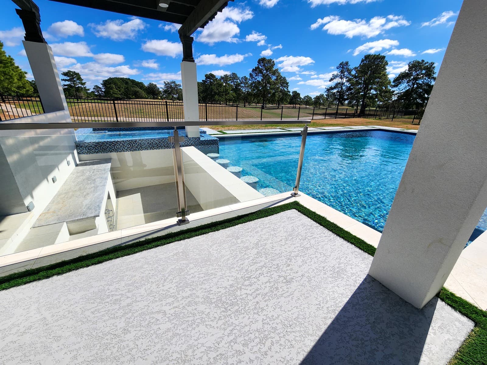Custom pool in Cypress, TX. A swim-up bar with round stools set in the water, a sunken lounge behind a glass railing, white pergola columns and a pale deck edged in strips of turf, open land beyond the fence.