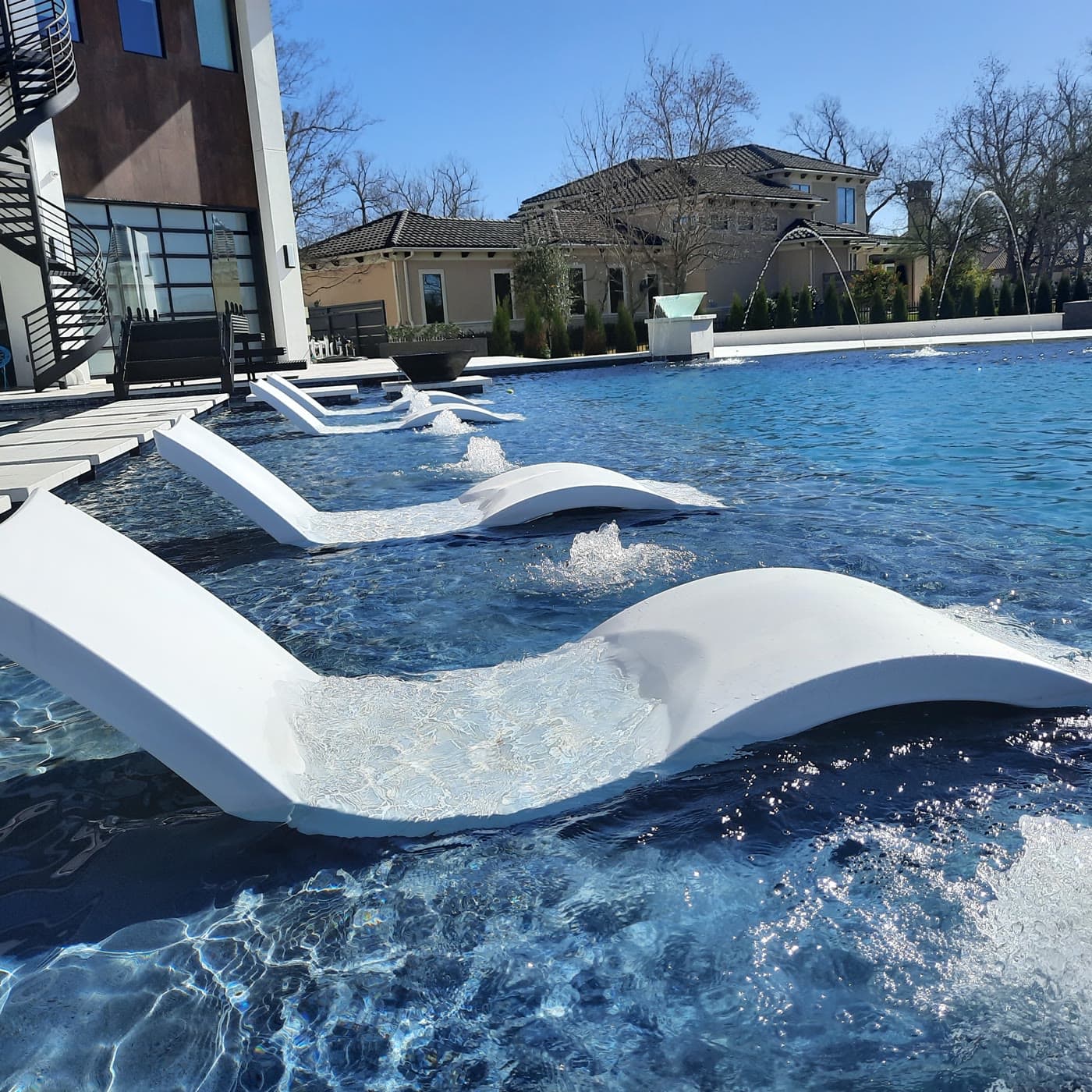 Custom pool in Richmond, TX. White loungers set into the shallow end in a receding row, bubblers churning under each one, the water reading deep sapphire against them.