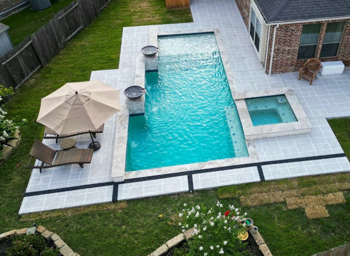 Custom pool in Spring, TX. Seen from above: a geometric pool with two scupper bowls on stacked-stone pedestals along one side, a square raised spa, and a spray deck scored in a travertine-look pattern with dark pebble bands.