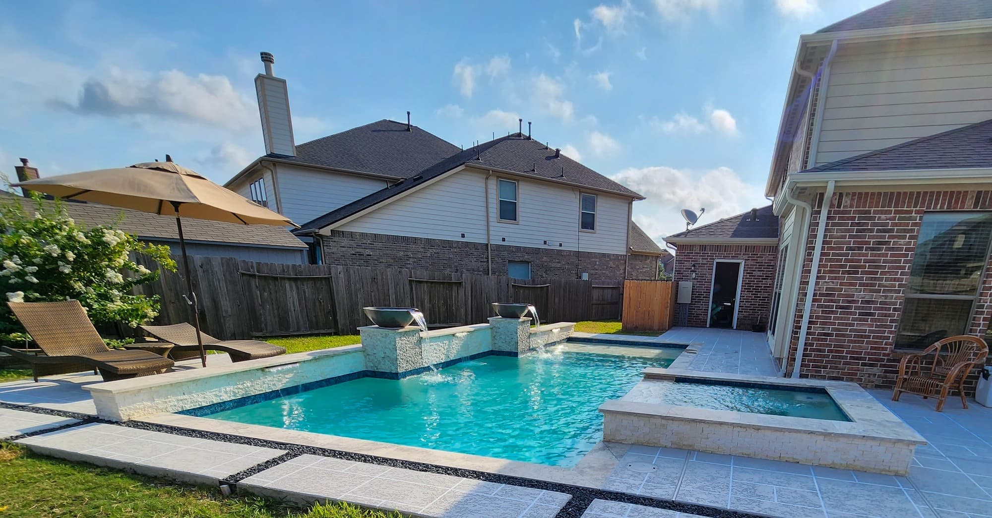 Custom pool in Spring, TX. The finished yard seen from the lawn: a geometric pool, two scupper bowls pouring from a raised stacked-stone wall, a square raised spa faced in stacked stone and the brick house behind.