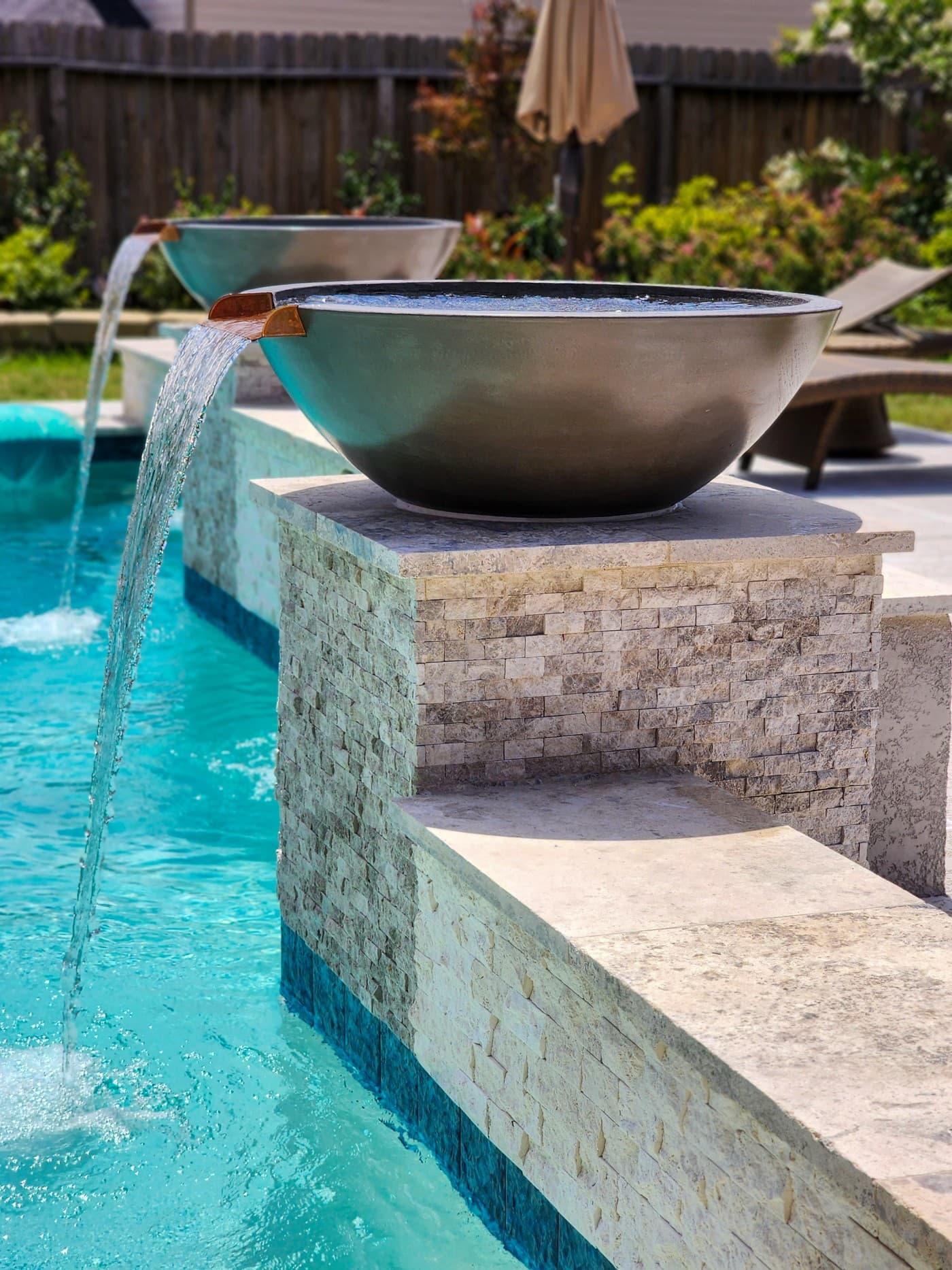 Custom pool in Spring, TX. A bronze scupper bowl on a stacked-stone pedestal, water pouring from its copper spout into turquoise water, a second bowl behind it.