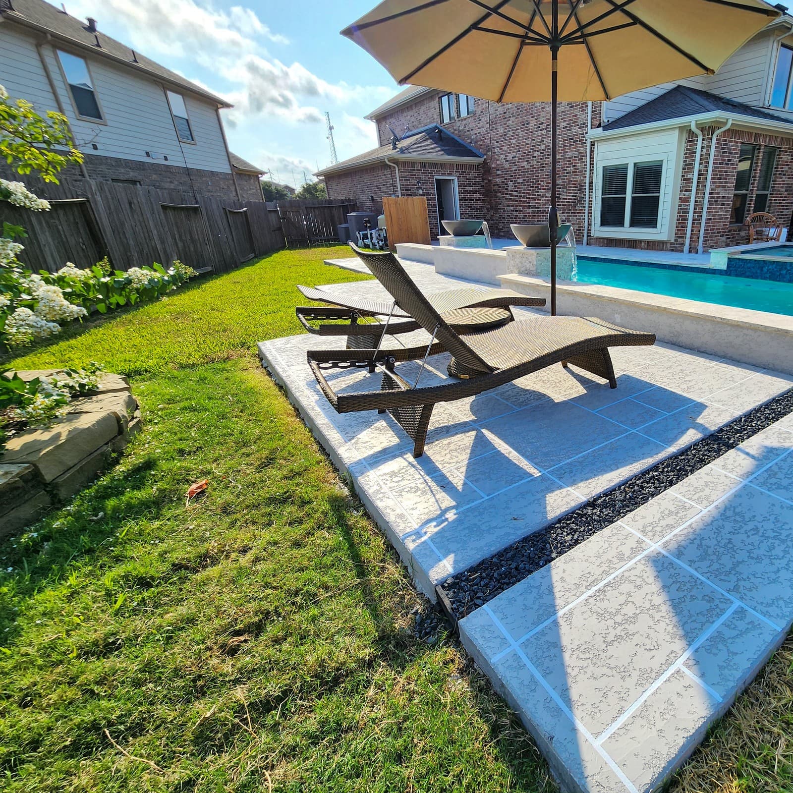 Custom pool in Spring, TX. Two wicker loungers on a spray deck finished in a travertine-look pattern and edged with a dark pebble band, the lawn beside it and the pool's raised wall behind.