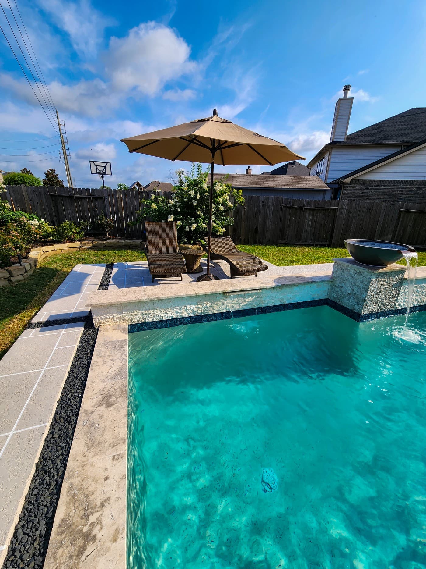 Custom pool in Spring, TX. A tan umbrella over two wicker loungers at the far end of the spray deck, the pool in the foreground and a scupper bowl pouring in the corner.