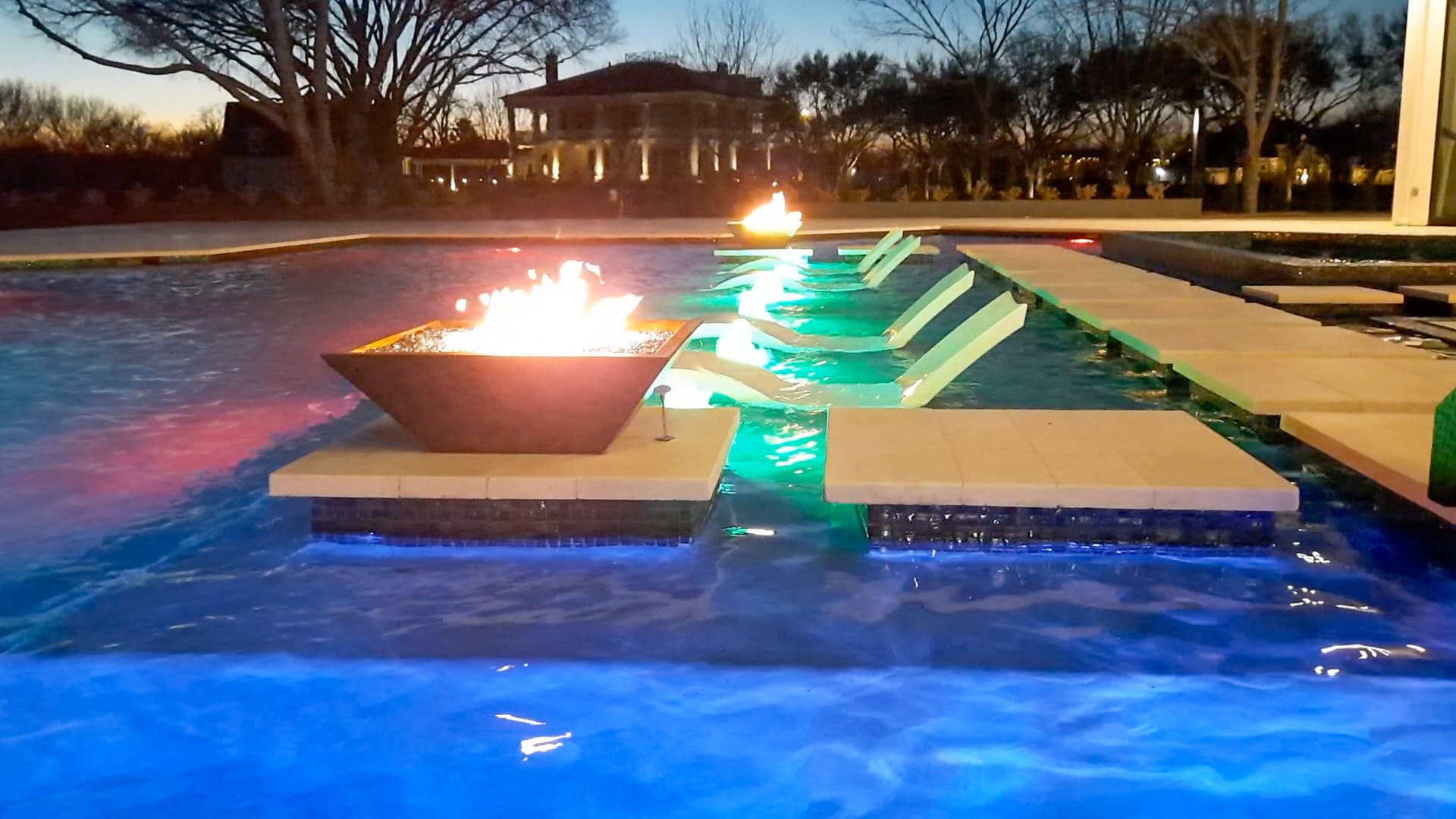 Custom pool in Sugar Land, TX. At dusk, a square fire bowl burns on its pedestal in the pool, the in-water loungers glow green under the LED lights, the water turns electric blue and a house is lit behind the bare trees.