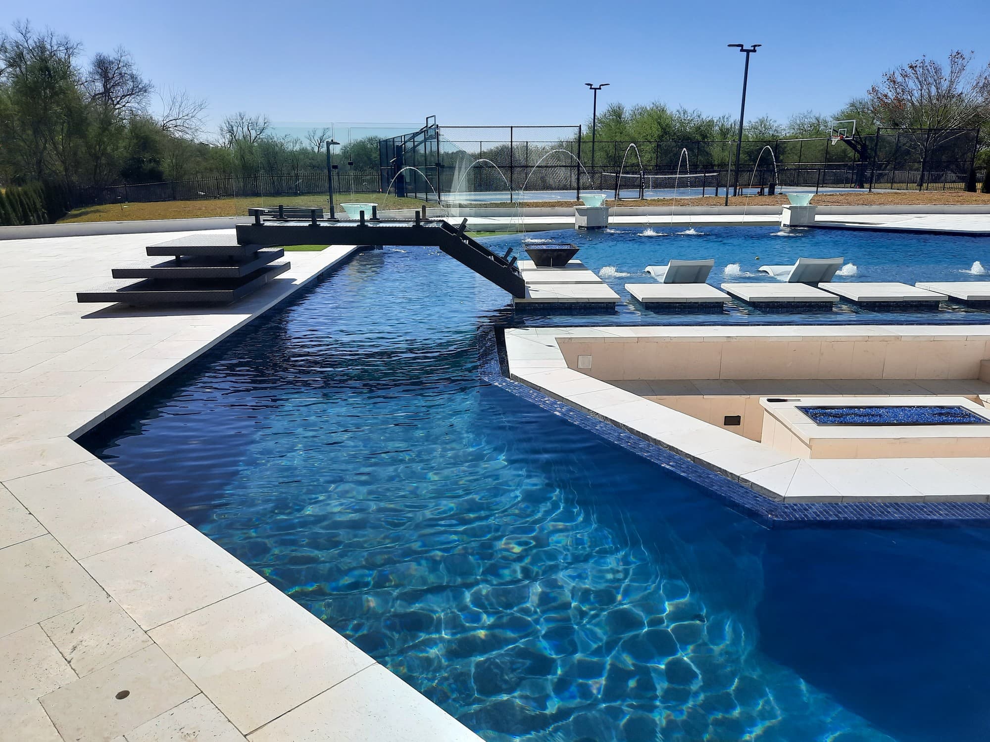 Custom pool in Sugar Land, TX. A steel bridge with a railing crosses a geometric pool of deep-blue water, deck jets arc over the shelf beyond it, square fire bowls stand at the far corners and a sport court sits behind the fence.