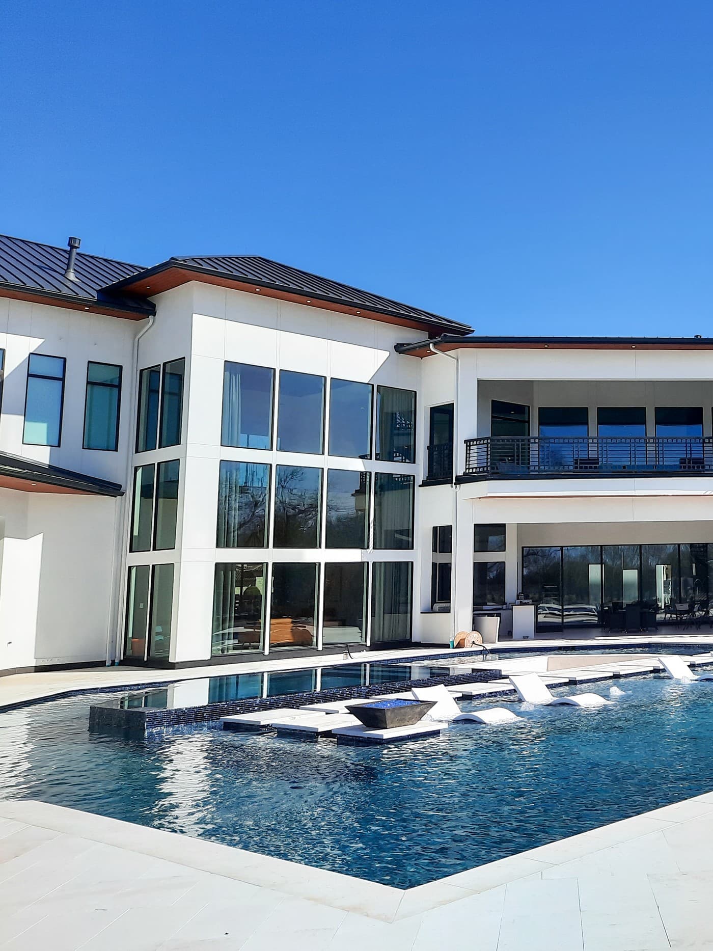 Custom pool in Sugar Land, TX. A two-story wall of glass on a white modern house rises over the geometric pool, with the fire bowl and the in-water loungers reflected in the dark-blue water at its base.