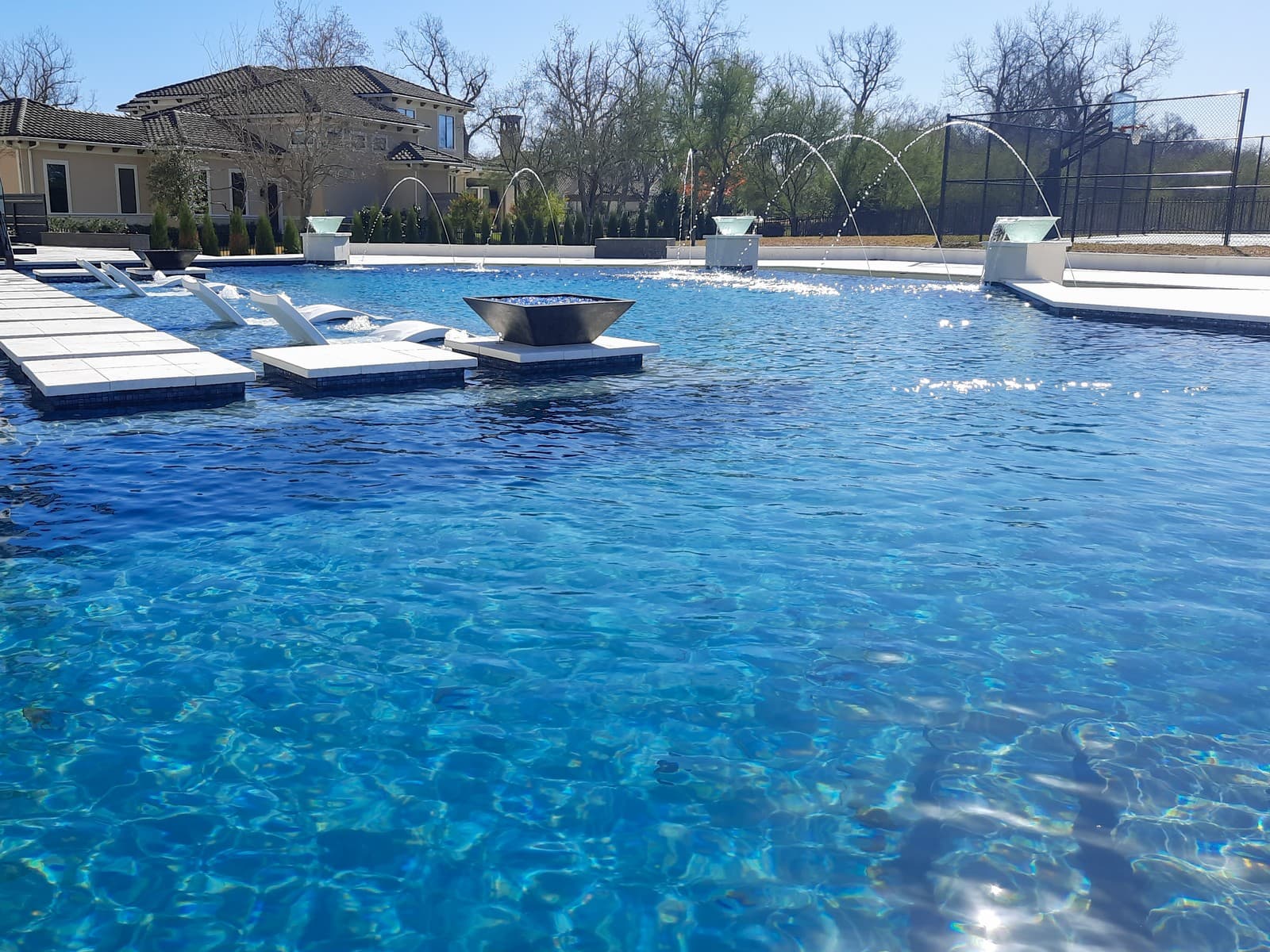 Custom pool in Sugar Land, TX. White loungers set into the shelf of a geometric pool, a square fire bowl on a pedestal in the water beside them, deck jets arcing over the shallows and a basketball court behind the far fence.