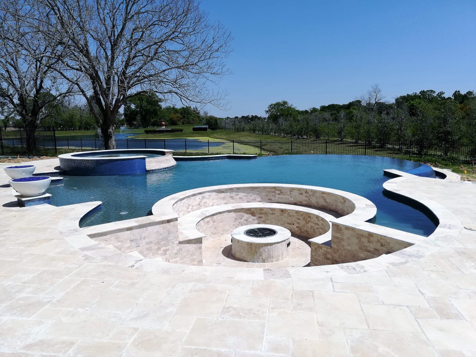 Custom pool in Sugar Land, TX. A sunken fire-pit lounge in travertine sits at the water's edge, a round mosaic spa behind it, and the infinity edge runs out toward a lagoon at the back of the property.