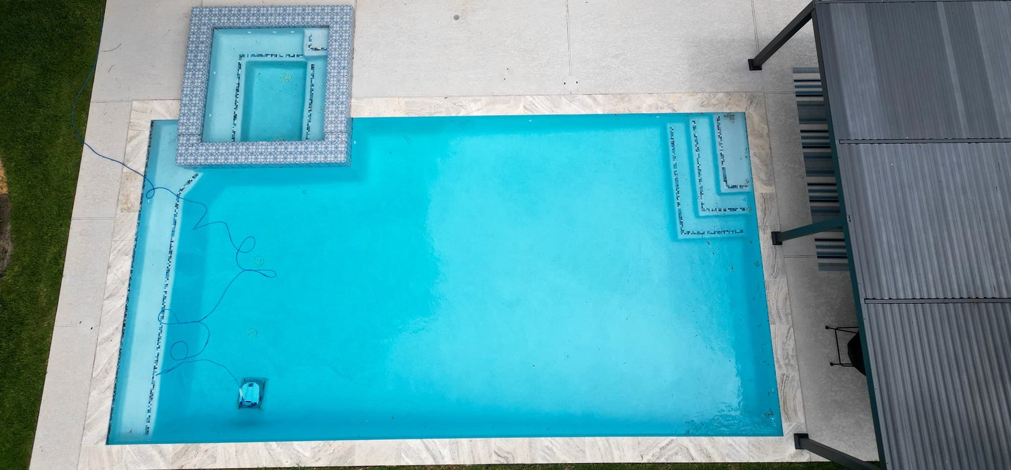 Custom pool in The Woodlands, TX. Seen from above: a geometric pool with a square spa in blue-and-white patterned tile at one corner, a light deck around it and a pergola along one side.