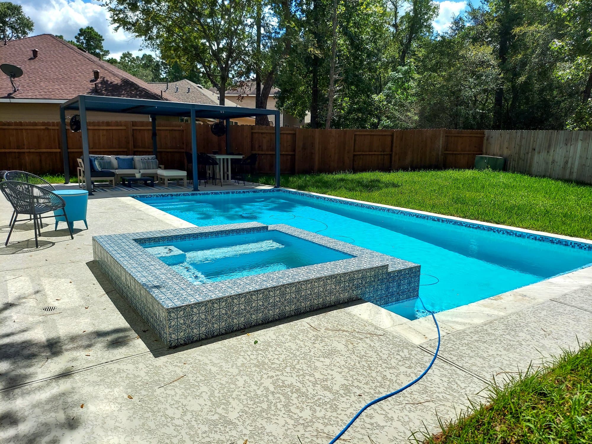Custom pool in The Woodlands, TX. A geometric pool with a square raised spa wrapped in blue-and-white patterned tile, a dark pergola with lounge seating at the far end and tall pines behind the wood fence.