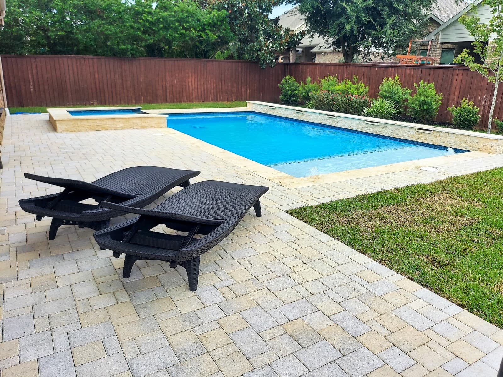 Custom pool in The Woodlands, TX. Two black wicker loungers on a grey paver deck beside a geometric pool, a raised stacked-stone wall with shrubs behind it and the spa at the far corner.