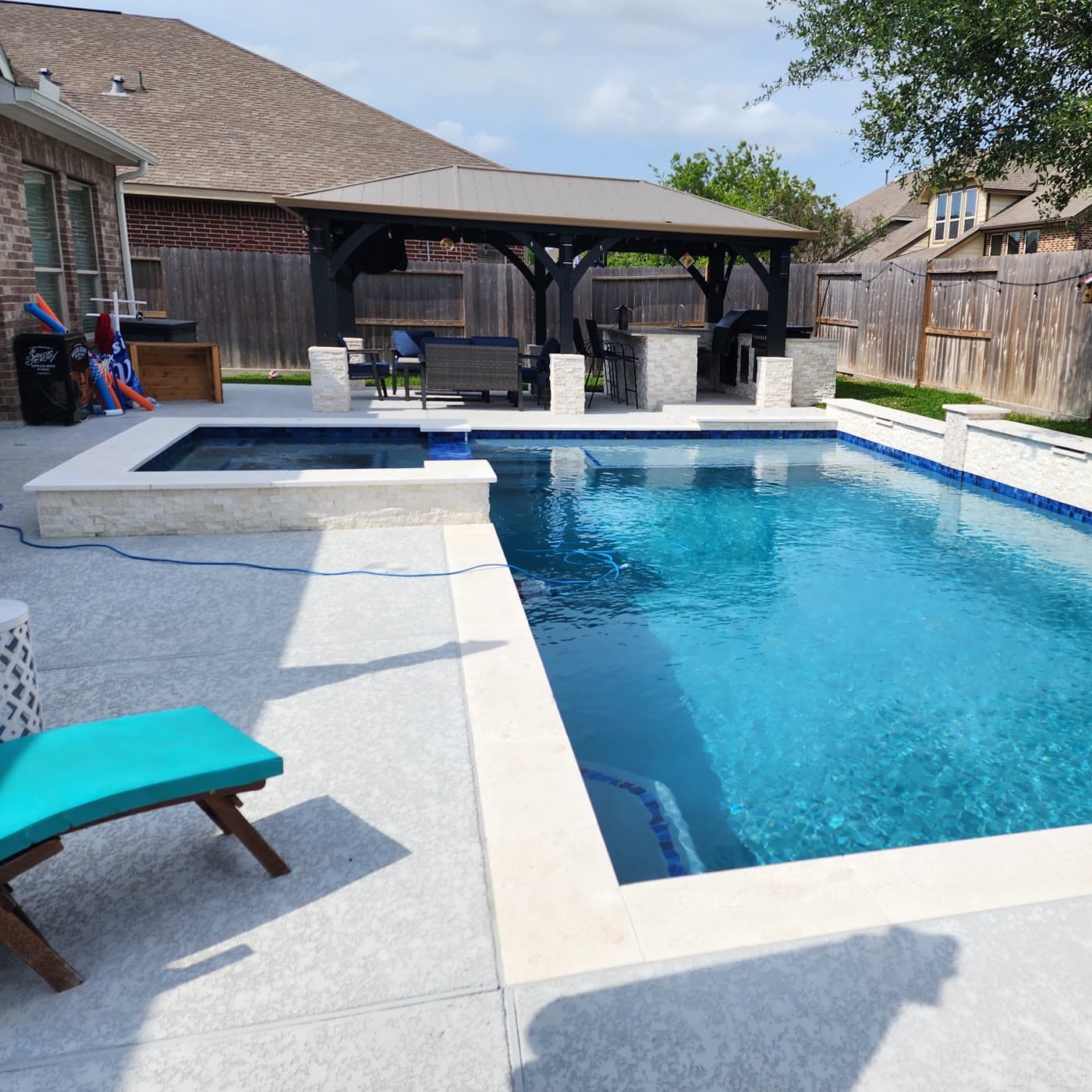 Custom pool in The Woodlands, TX. A rectangular pool and raised spa in white stacked stone, with a dark timber pergola and an outdoor kitchen along the back of the yard.