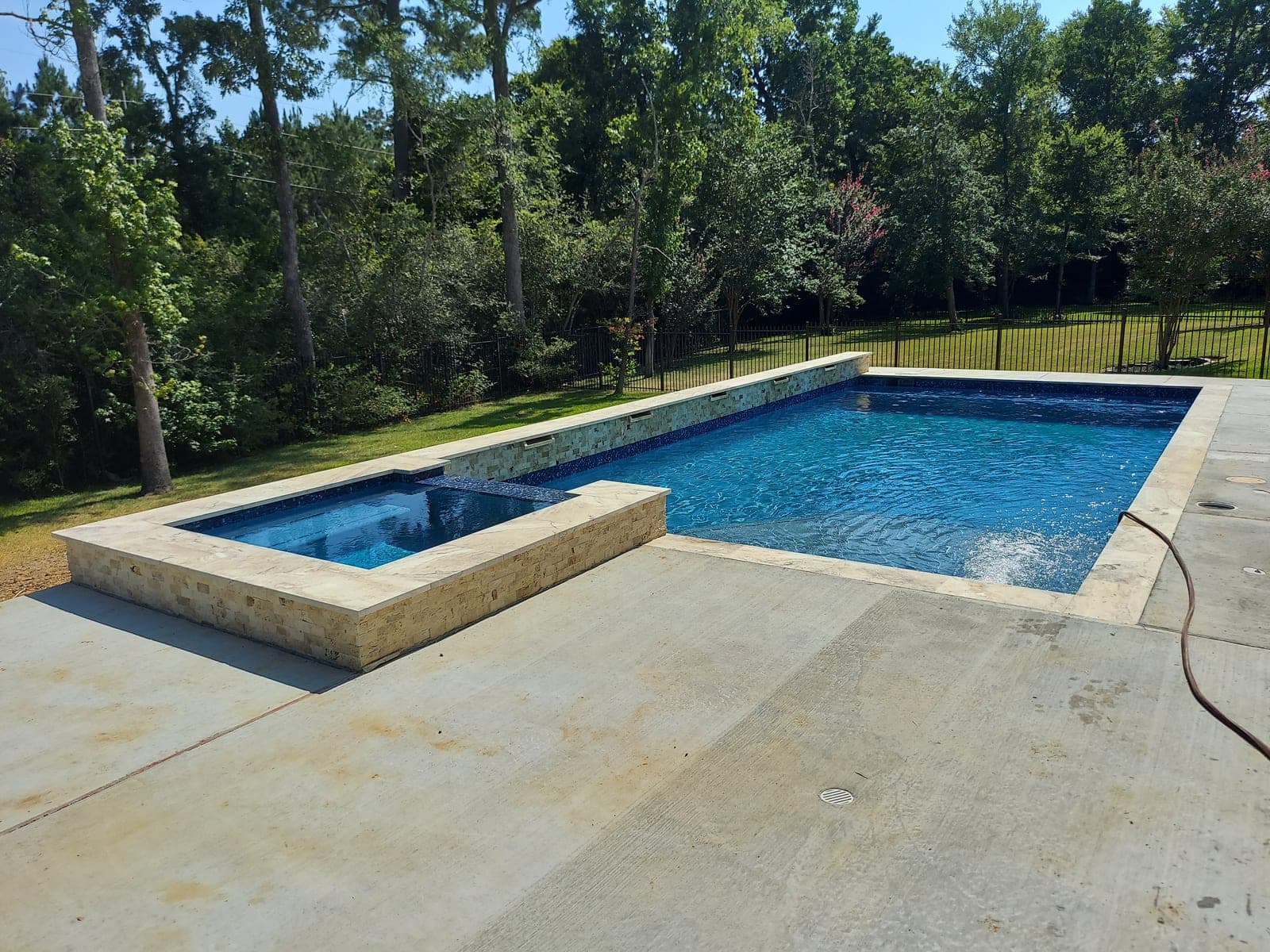 Custom pool in The Woodlands, TX. A raised spa clad in stacked travertine spills its water over the wall into deep blue below, a wrought-iron fence and a dense line of pines standing behind it.