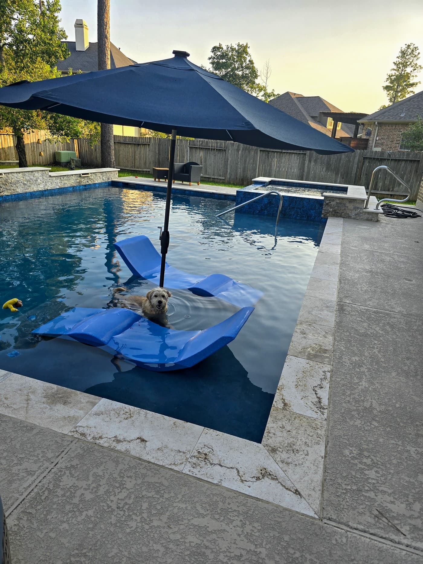Custom pool in Tomball, TX. At dusk, two blue floating loungers in the shallow end under a navy umbrella, a small dog aboard one, a raised spa in blue mosaic and a stone wall behind the dark water.