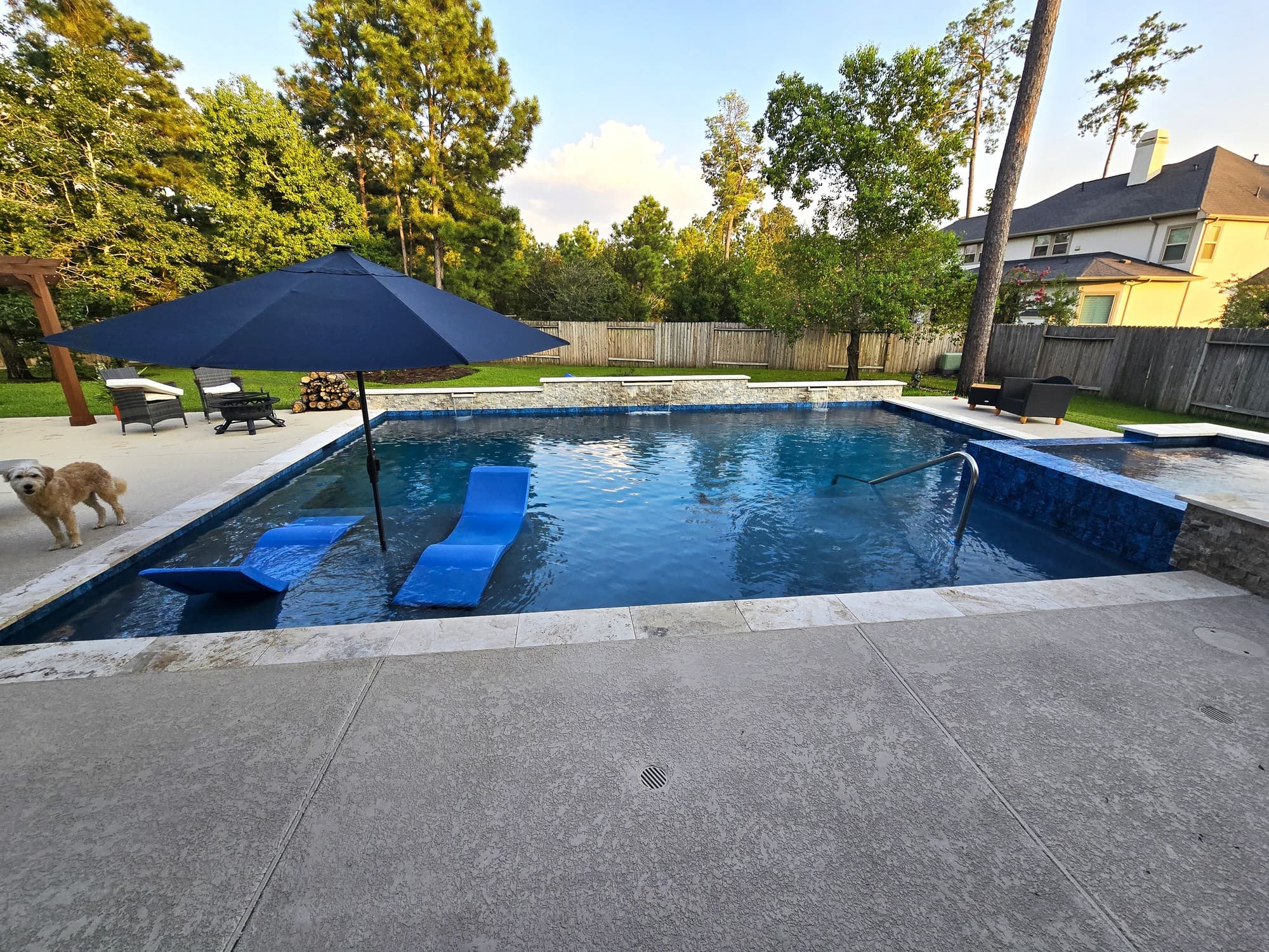Custom pool in Tomball, TX. The whole yard at dusk: the pool with two blue floating loungers under an umbrella, a raised stone wall with planters, the spa at right and a pergola with chairs at left, pines behind the fence.
