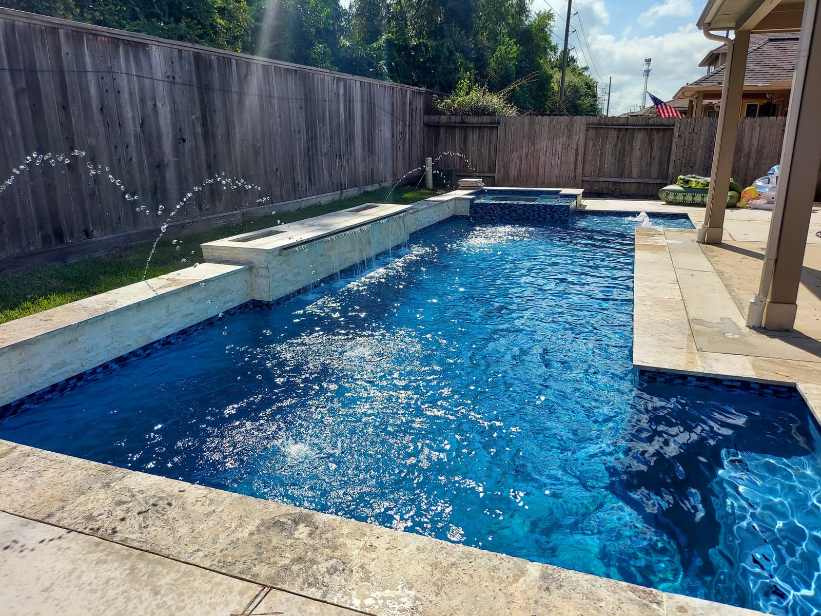 Custom pool in Tomball, TX. A geometric pool in deep blue with two sheer descents pouring from a raised wall of light stacked stone, deck jets arcing over the water and a spa at the far end.