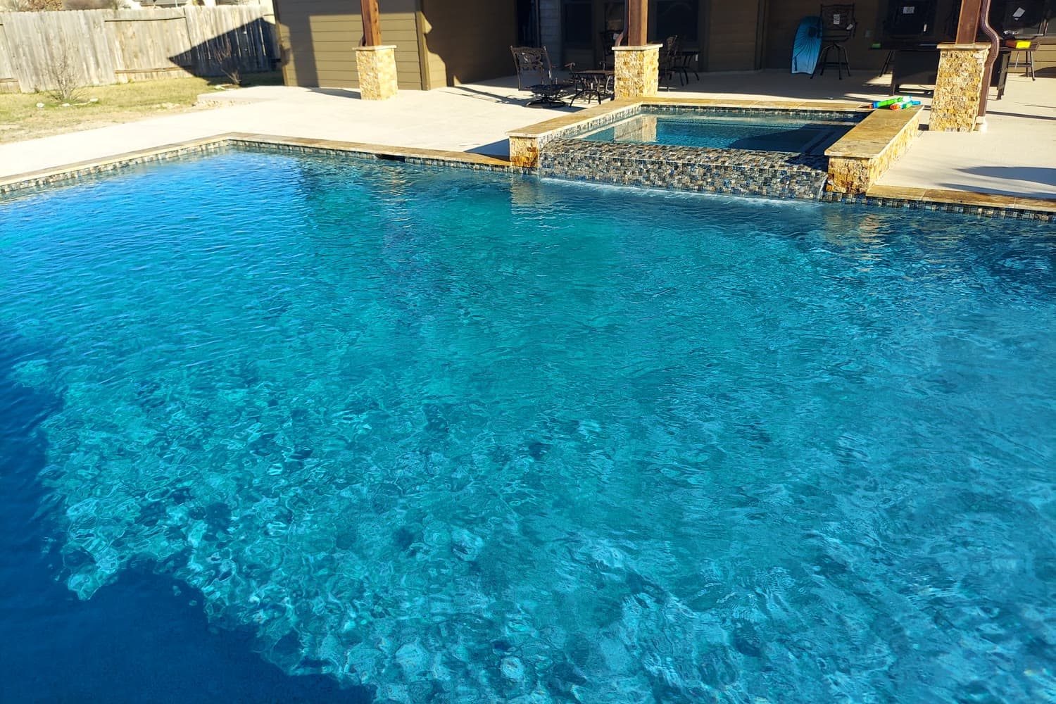 Custom pool in Tomball, TX. Low winter sun: the raised spa spills over a face of glass mosaic into the deep end, travertine coping runs along the edge, and the light breaks across the floor in a net.