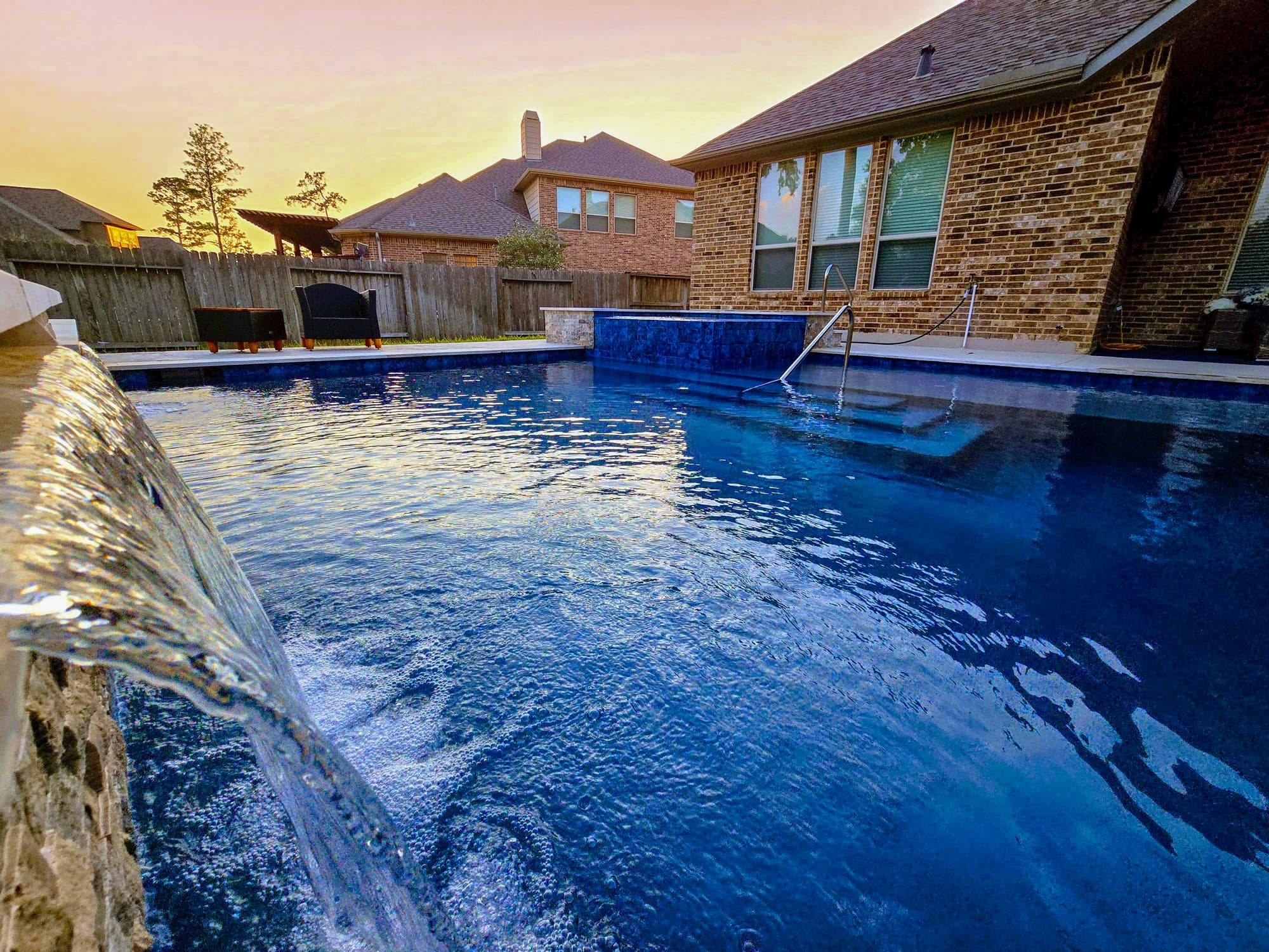 Custom pool in Tomball, TX. At sunset, a sheer descent pours off a stone ledge into deep-blue water, a raised spa faced in blue mosaic at the far end and the brick house behind it.