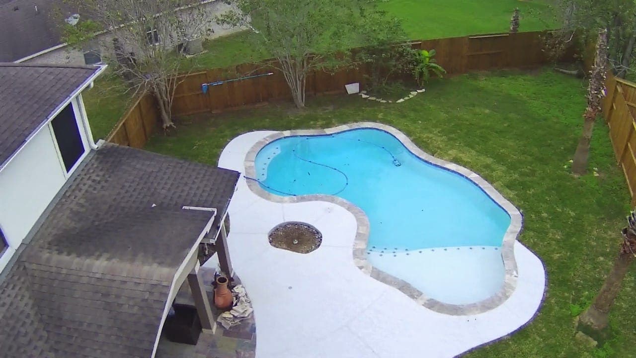 Inground pool in Missouri City, TX. Seen from above: a freeform shape in white plaster, the water reading pale turquoise over it, a broad white deck and the covered patio at the corner of the yard.