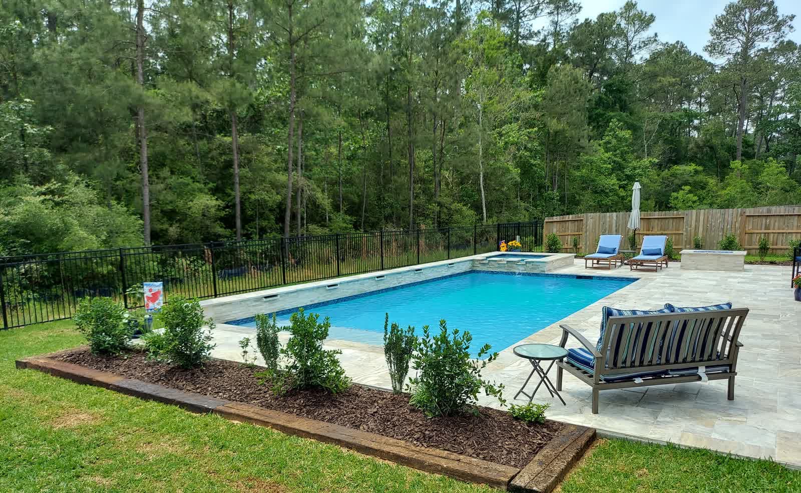 Piscina enterrada en Tomball, TX. Una piscina rectangular con spa integrado sobre deck de travertino, el bosque de pinos levantándose detrás de la valla y un macizo recién plantado en el borde cercano.